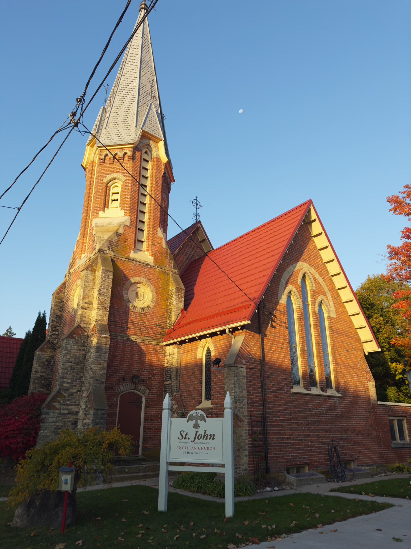 Photograph of St. John Anglican Church, a brick church with a tall steeple, red roof, Gothic windows, and a sign in front. The sky is clear with the moon visible.