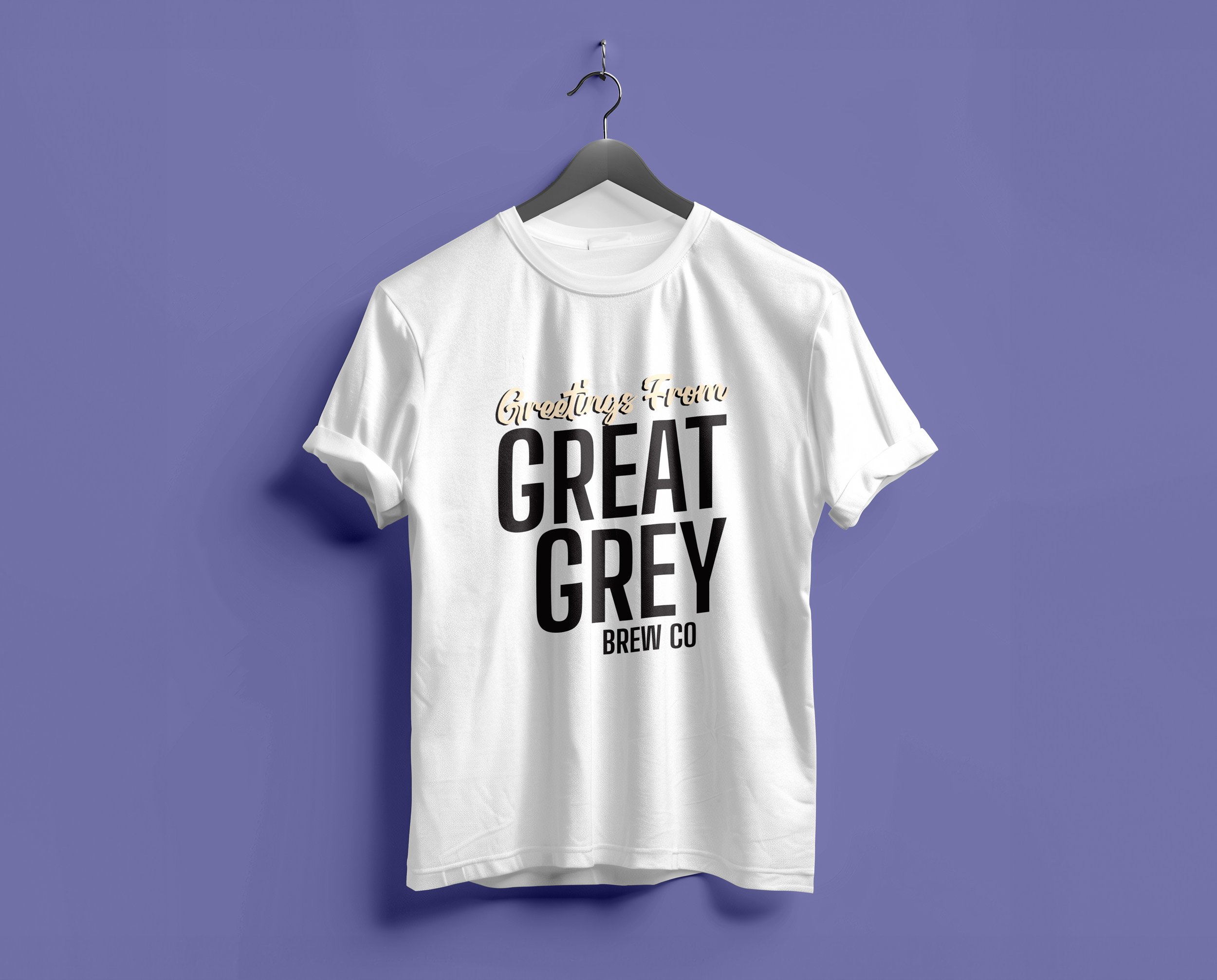 White T-shirt hanging on a gray hanger against a purple background. The T-shirt has text that reads 'Greetings from GREAT GREY BREW CO' with 'GREAT GREY' in large black letters and the other text in smaller, cursive gold font.