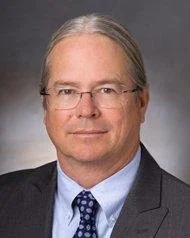 A middle-aged man with gray hair, glasses, wearing a dark suit, blue shirt, and patterned tie, against a gray background.