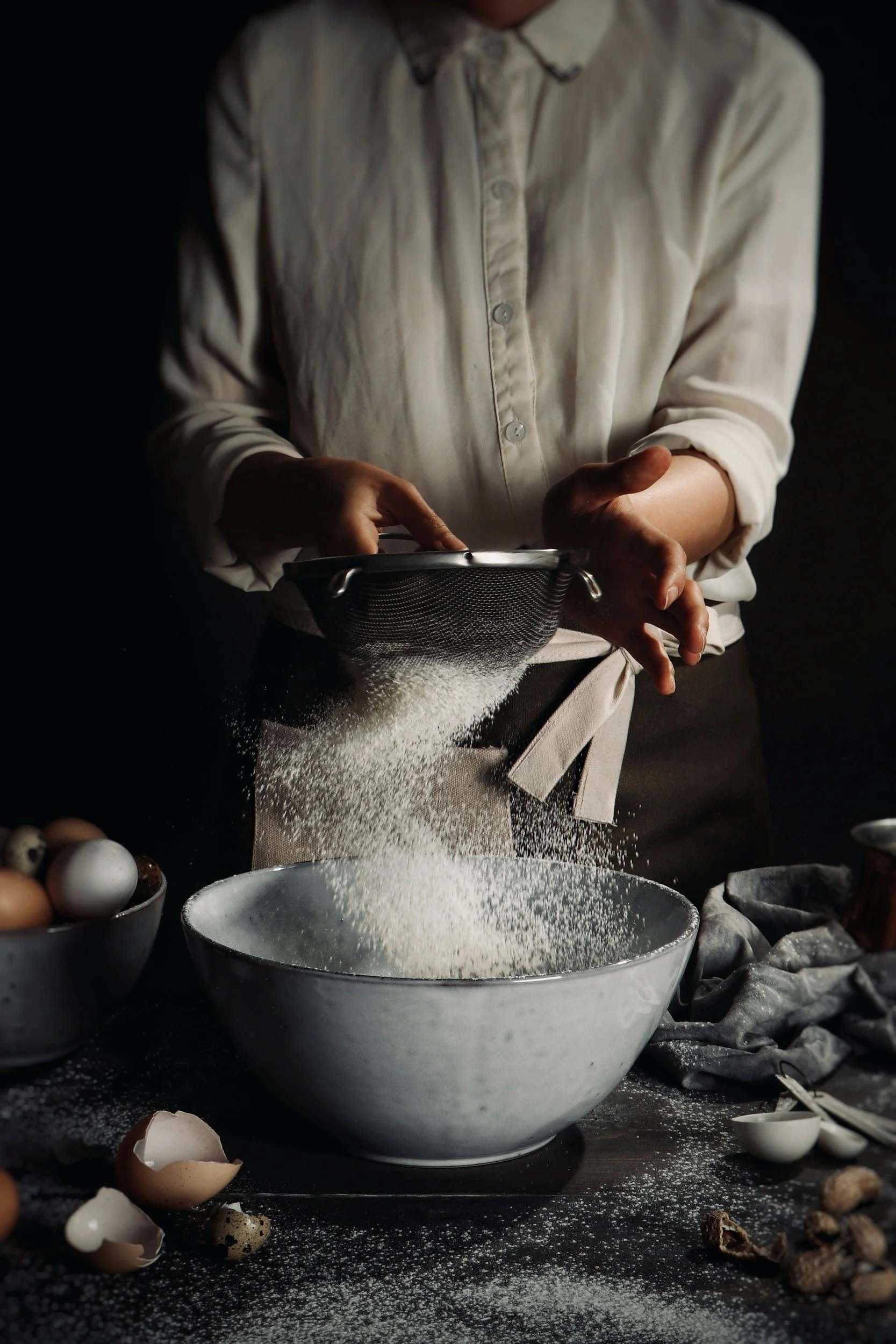 Person sifting flour into a mixing bowl in a dark kitchen.
