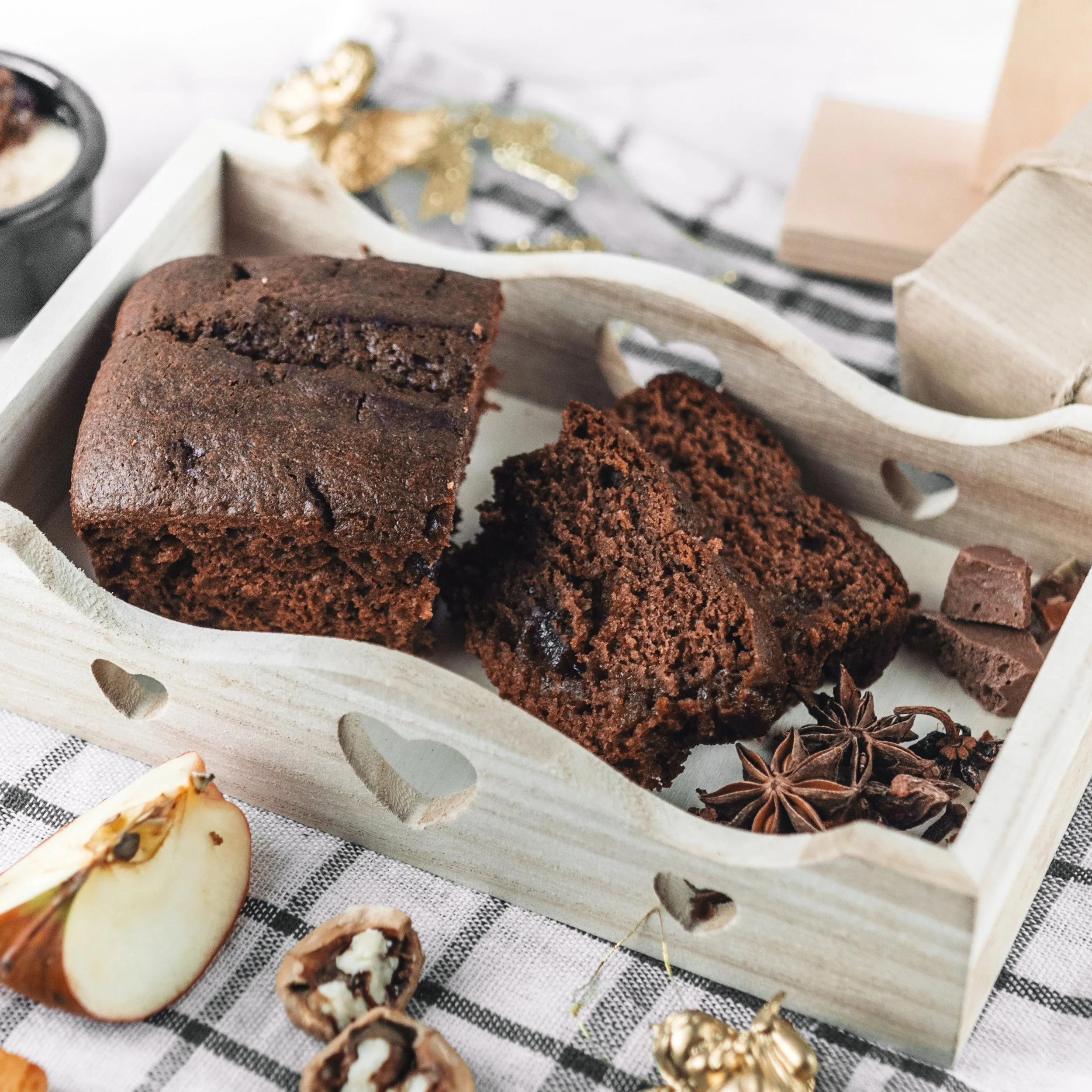Chocolate cake slices with star anise in a wooden tray, apple and walnuts on checkered cloth.