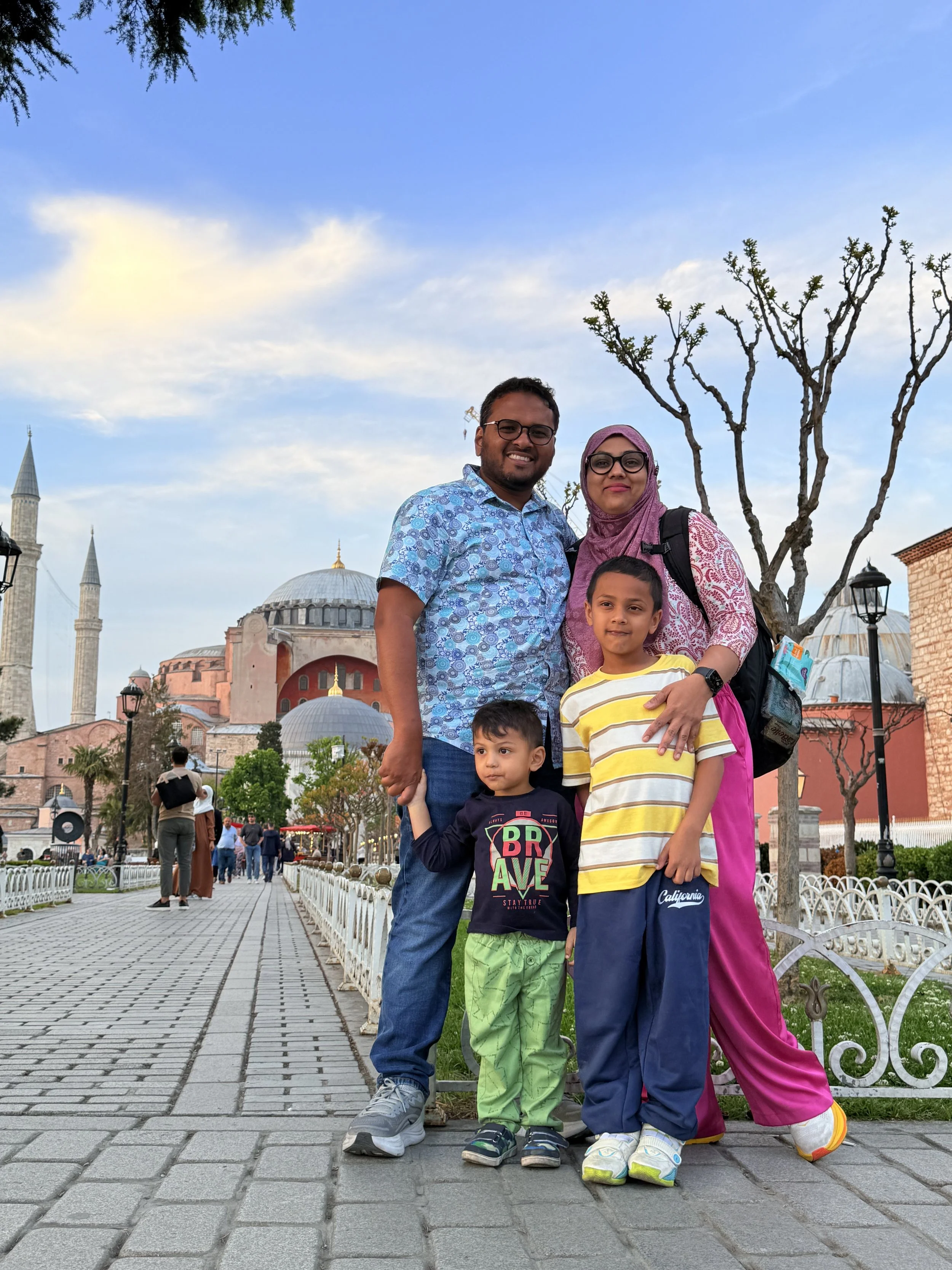 A family of five posing together on a sidewalk with historic architecture, including the Hagia Sophia in Istanbul, Turkey, in the background. The family includes a man, a woman wearing a hijab, and three children. The scene is during the daytime with a partly cloudy sky.
