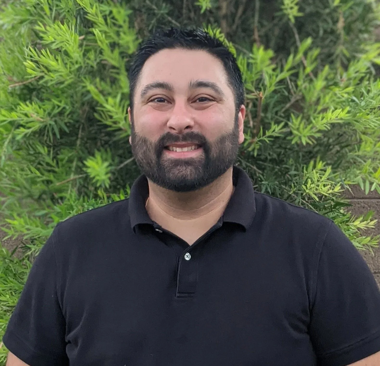 Smiling man with dark hair and a beard, wearing a black polo shirt, standing outdoors in front of green foliage.