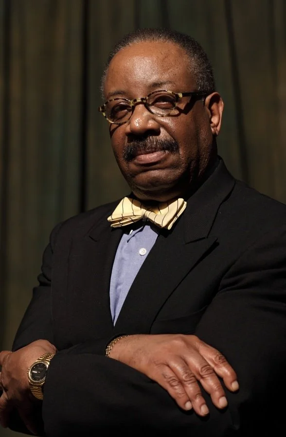 A well-dressed African American man with glasses, sporting a mustache, in a suit with a light blue shirt and a yellow bow tie, standing with arms crossed in front of a dark curtain.