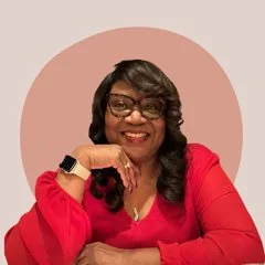 A woman with long dark hair and glasses, smiling and resting her chin on her hand, dressed in a red top, with a pink background.