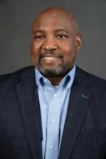 A portrait of a middle-aged African American man with a beard, smiling, wearing a dark blazer over a light blue dress shirt, against a gray background.