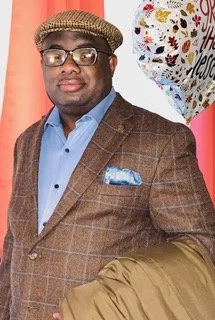 A man wearing glasses, a brown checked blazer, and a blue shirt, holding a gift bag, standing in front of colorful balloons.