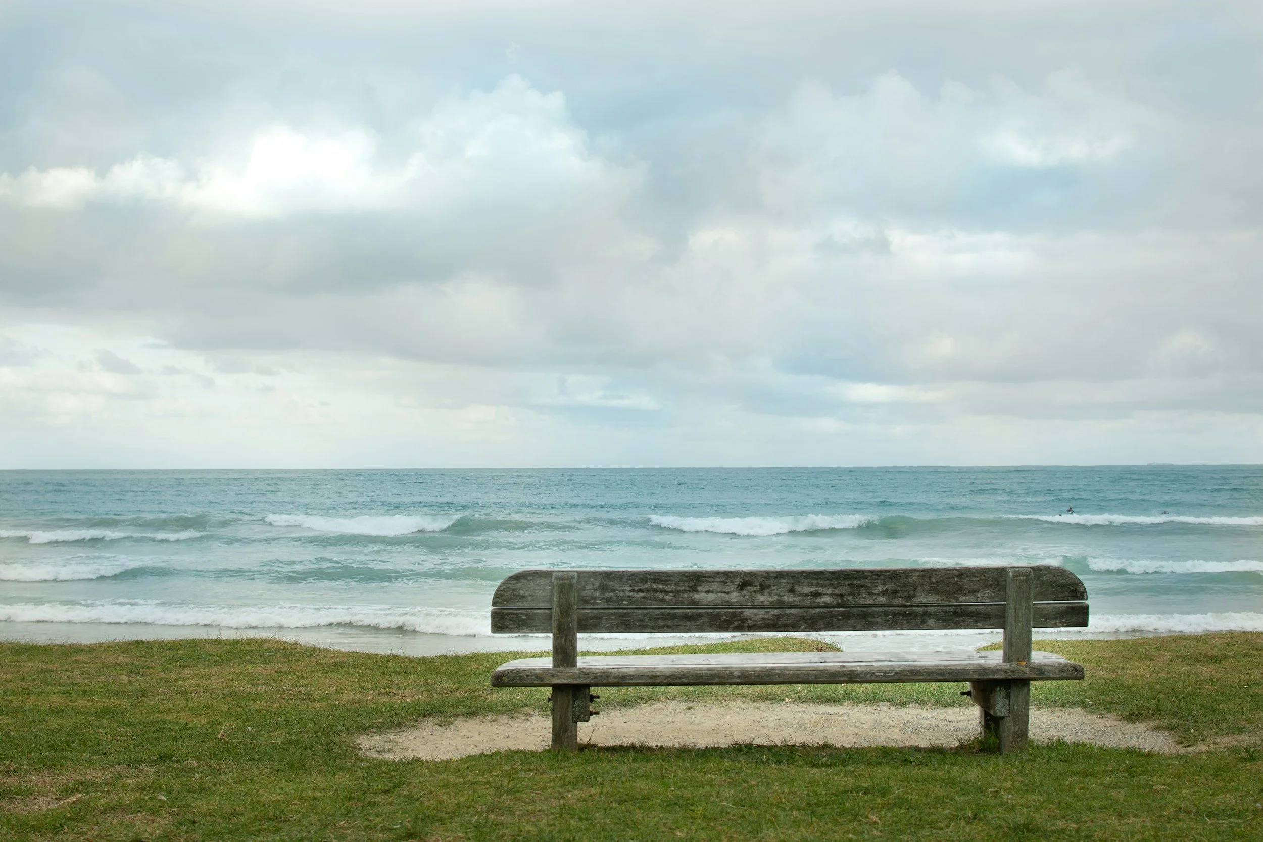 A wooden bench facing the ocean on a grassy beach with waves and cloudy sky