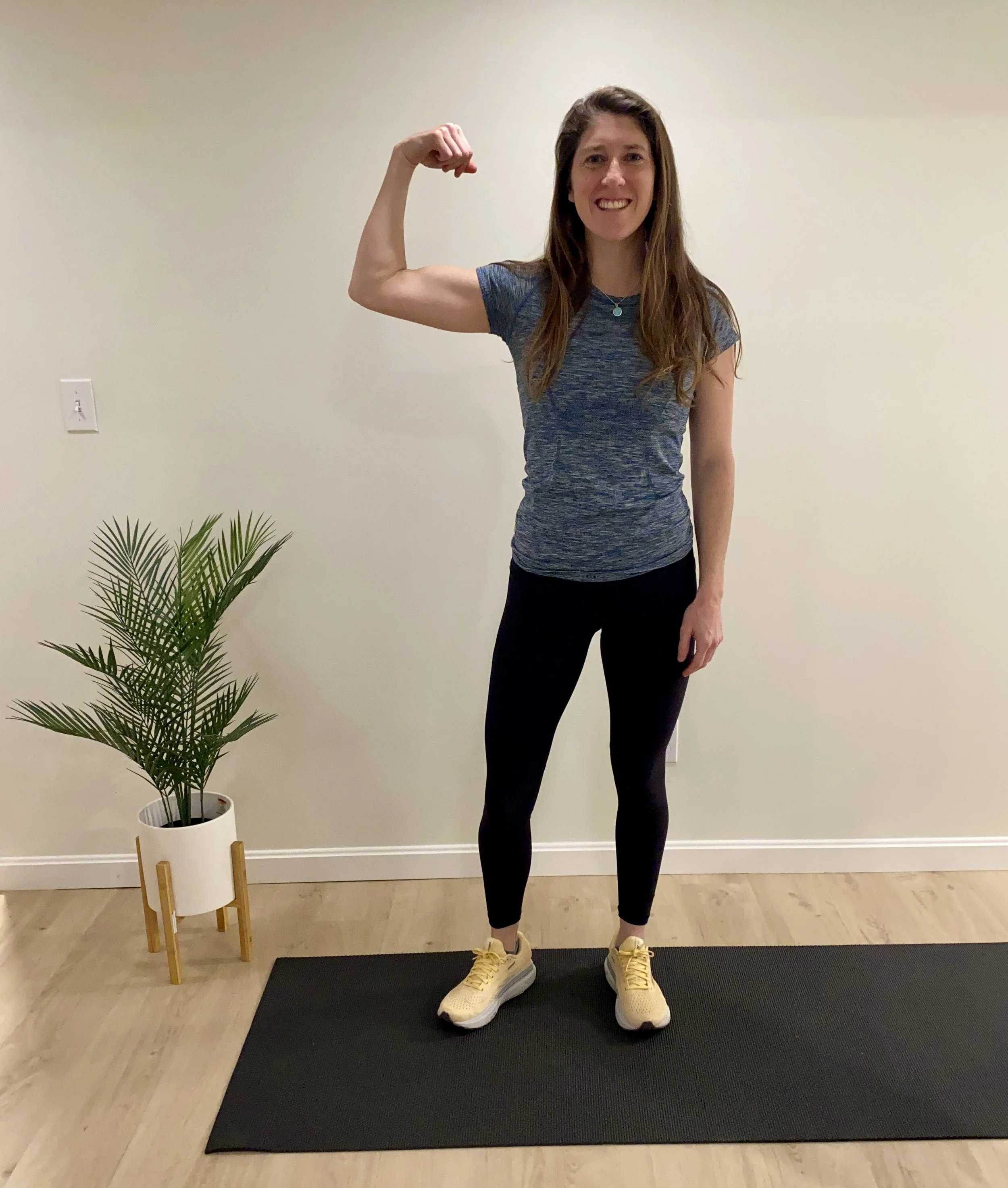 A woman smiling and flexing her bicep in a casual workout outfit, standing on a black exercise mat in a room with a potted plant and a plain wall.