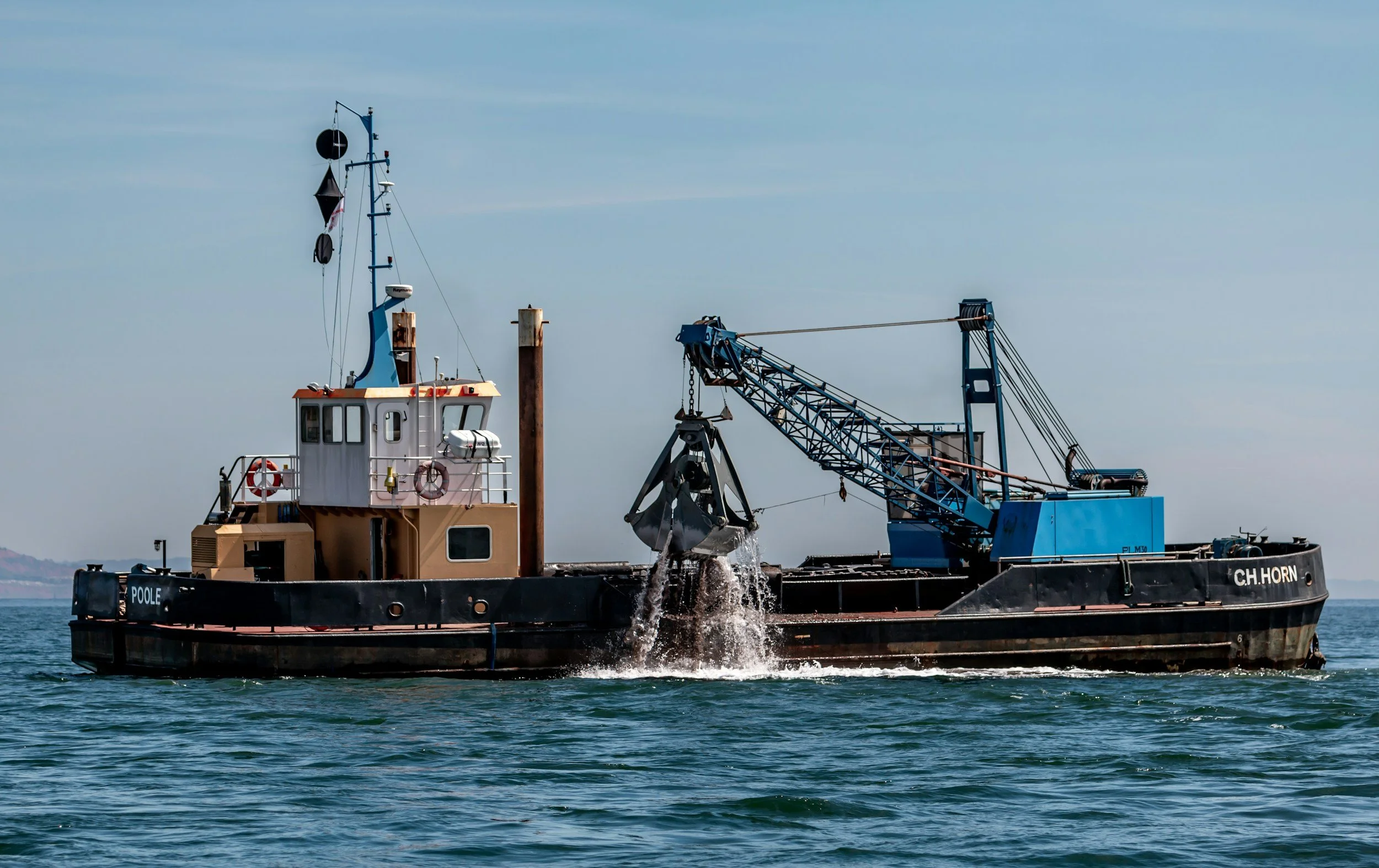 A boat on the water equipped with a crane for dredging or loading equipment, with a clear sky and calm sea.