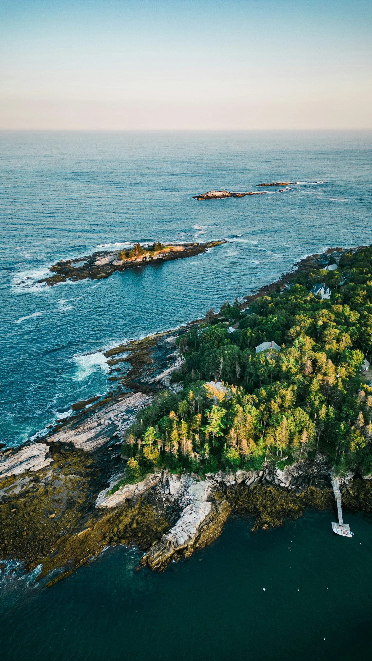 Aerial view of a rocky shoreline with trees and houses, extending into the ocean with small islands and a pier at the water's edge.