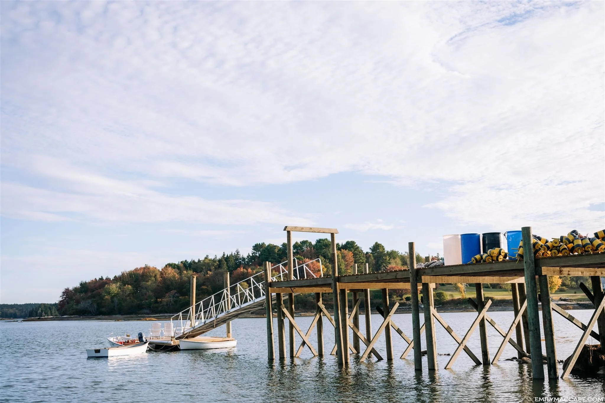 A wooden dock extending into a calm body of water with two small boats tied to it. The background features a tree-lined shore with trees showing fall colors and a partly cloudy sky.