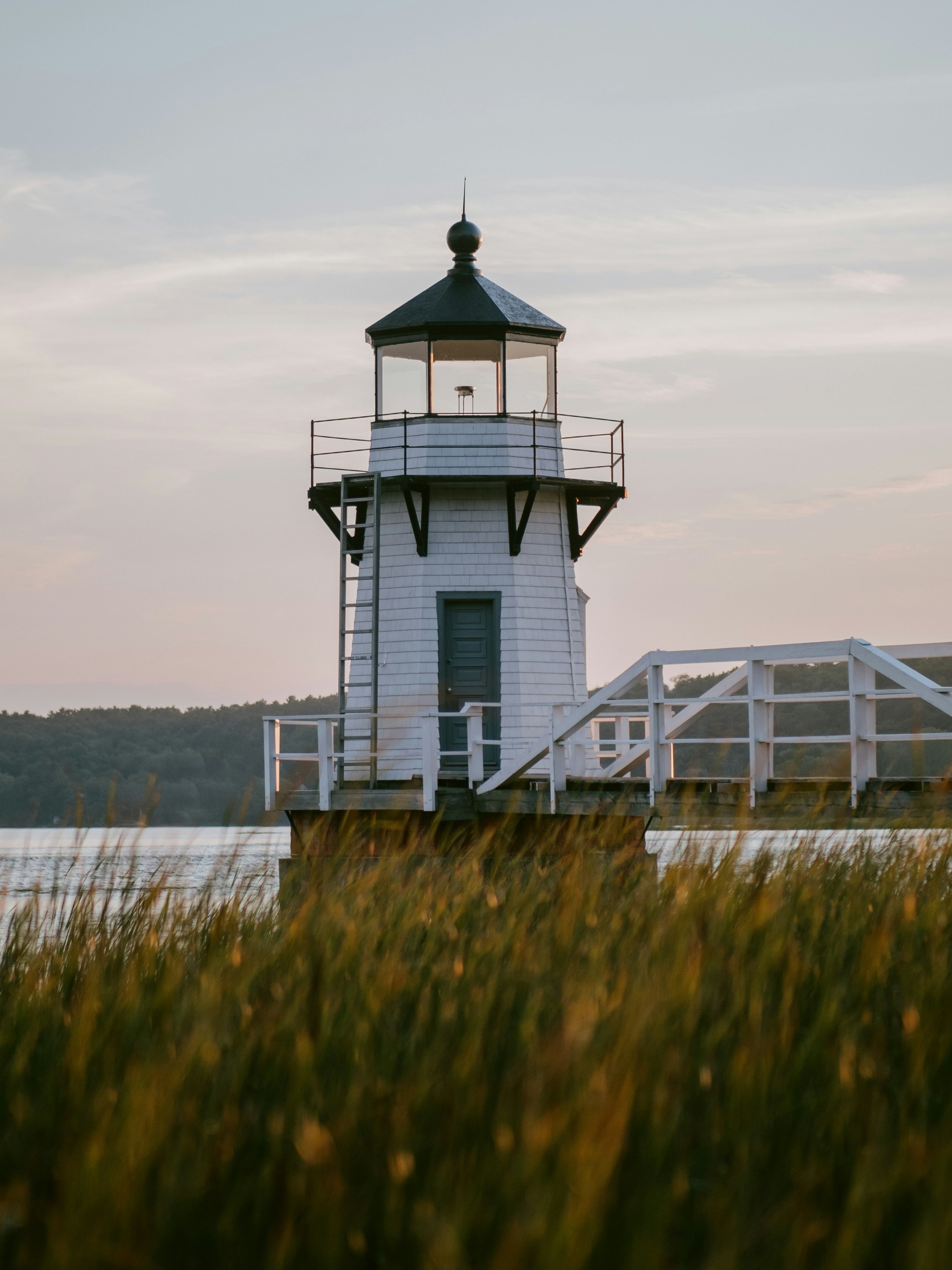 A white lighthouse with a black roof and railing, situated on a raised platform near water, with grass in the foreground and trees in the background.