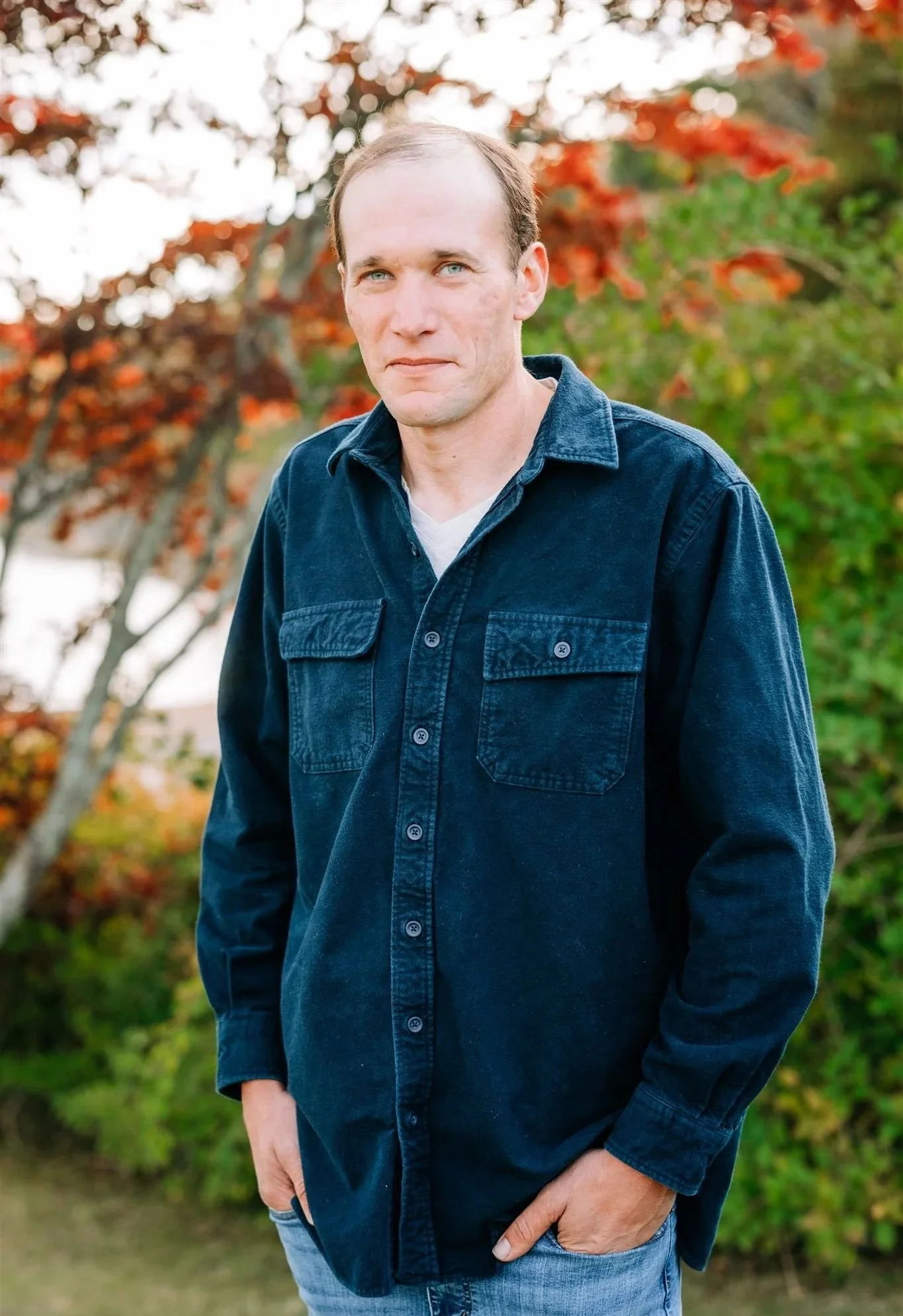 A man with fair skin, short light-colored hair, and blue eyes wearing a dark denim shirt and light blue jeans outdoors with autumn-colored trees in the background.