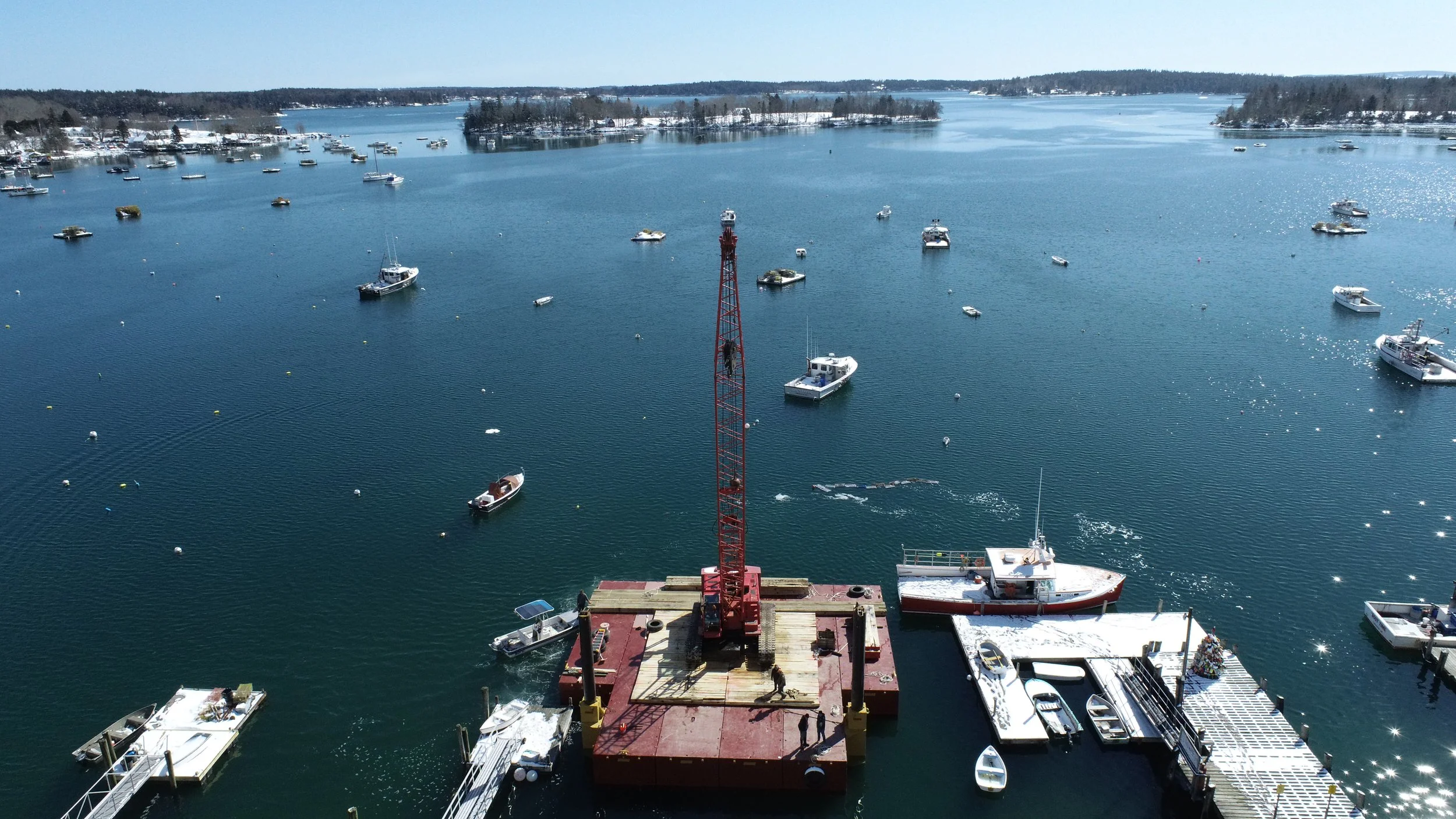 An aerial view of a harbor with multiple boats and yachts anchored on calm blue water, a red crane on a floating platform, and snow-covered land in the background.
