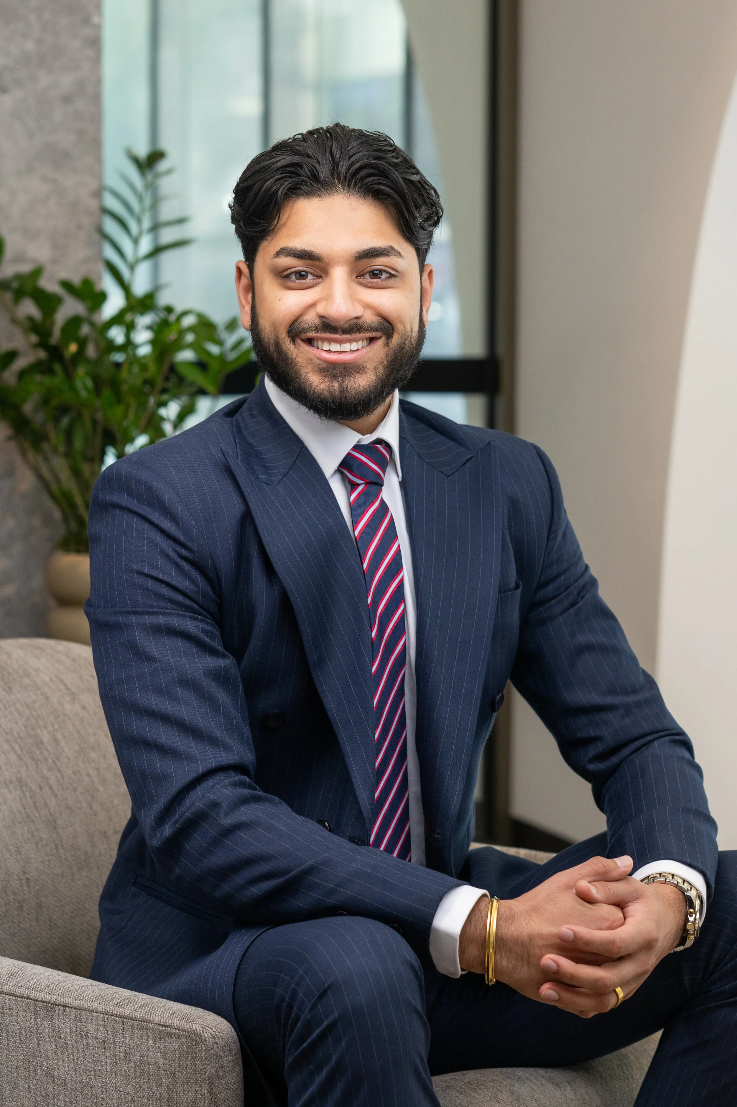 A smiling man with dark hair and a beard wearing a beige suit jacket, white shirt, and dark tie.