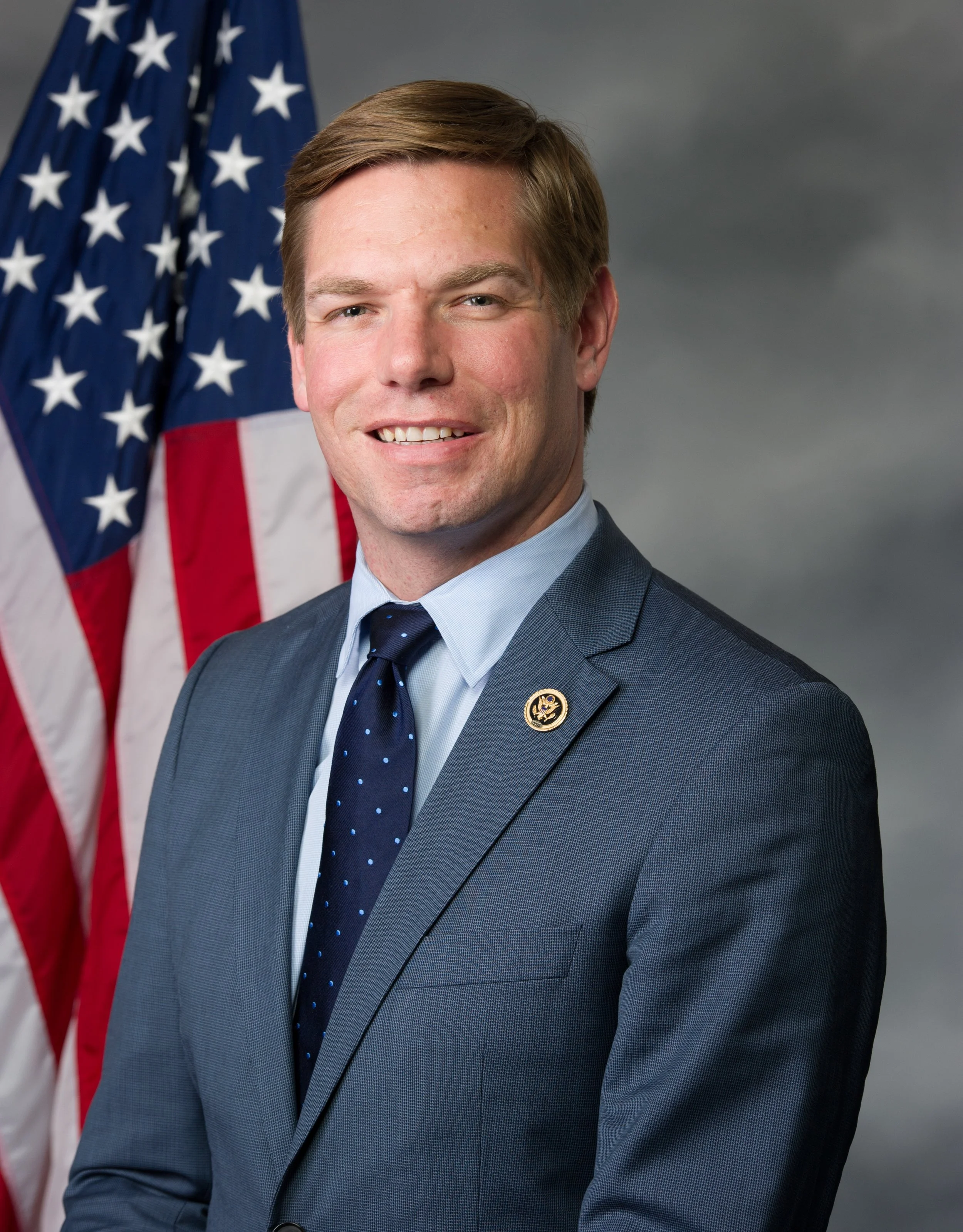 Photograph of a smiling man in a dark suit and tie, standing in front of a U.S. flag.