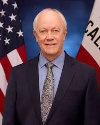 A man in a suit standing in front of American and California flags.