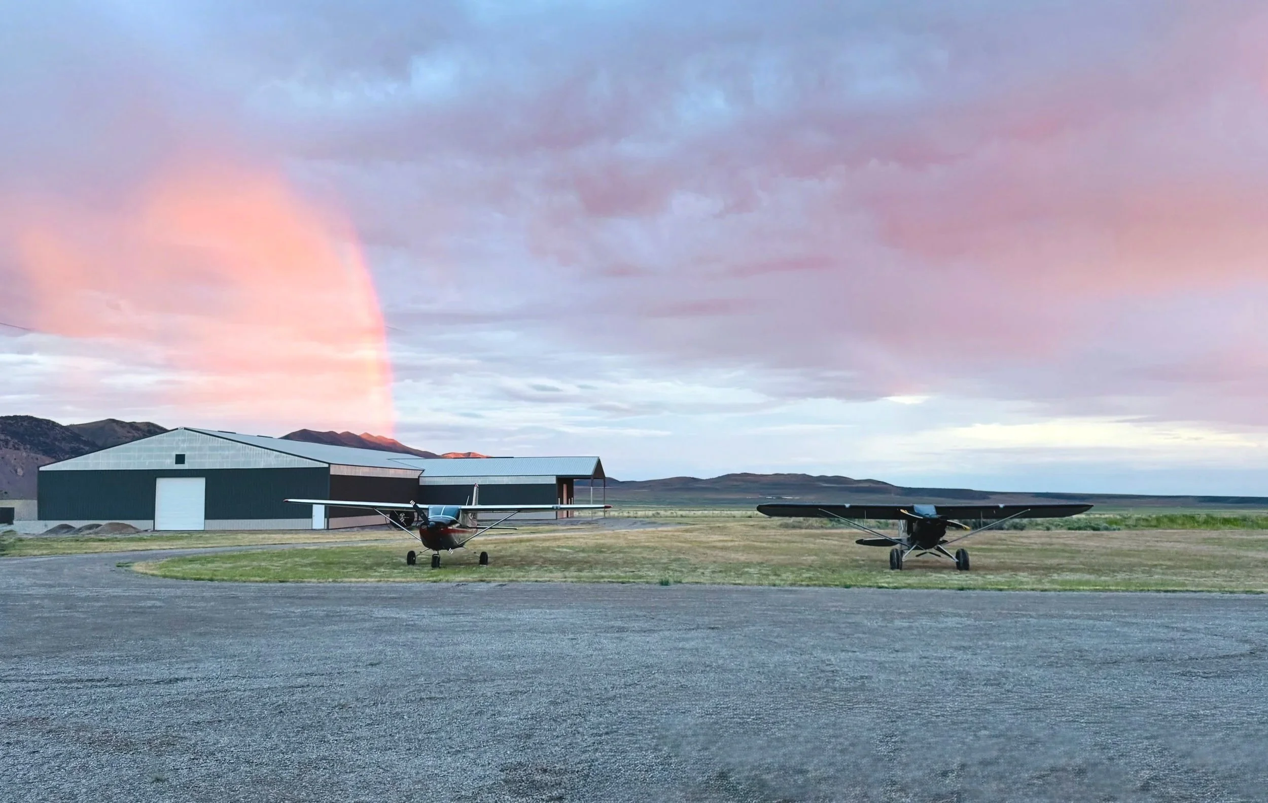 Two small airplanes parked on a grassy airstrip near a large hangar at sunset, with mountains and a colorful sky in the background.