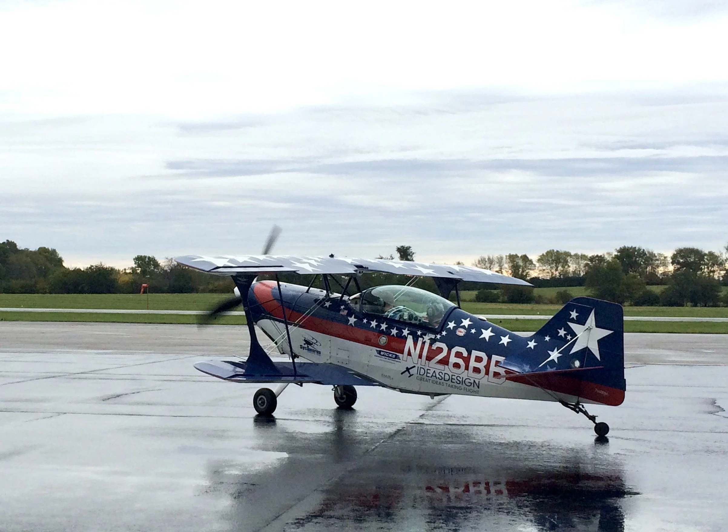 Small propeller aircraft with patriotic red, white, and blue star-spangled design on wet tarmac, overcast sky in background.
