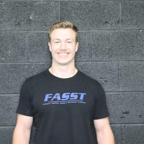 Young man smiling and standing in front of a black brick wall, wearing a black T-shirt with a blue logo that says 'FASST' and additional smaller text.