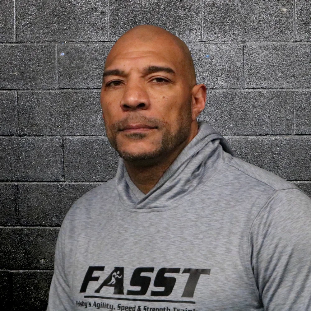 A man with a serious expression standing in front of a black brick wall, wearing a gray hoodie with the word 'FAST' printed on it.