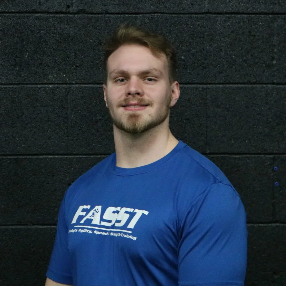 A young man with short red hair and a beard wearing a blue T-shirt with the logo 'FALST' standing against a black brick wall.