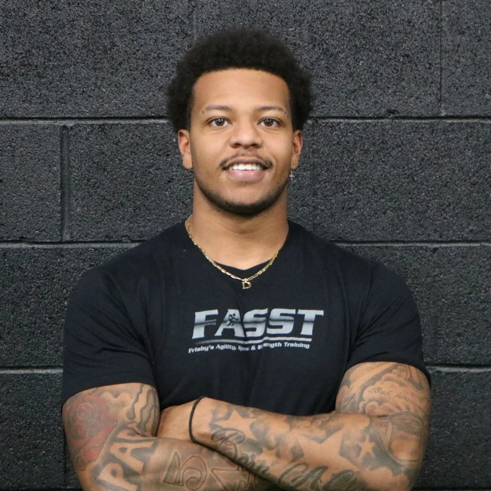 A young man with curly hair and tattoos on his arms, wearing a black T-shirt with a logo, standing against a black brick wall.