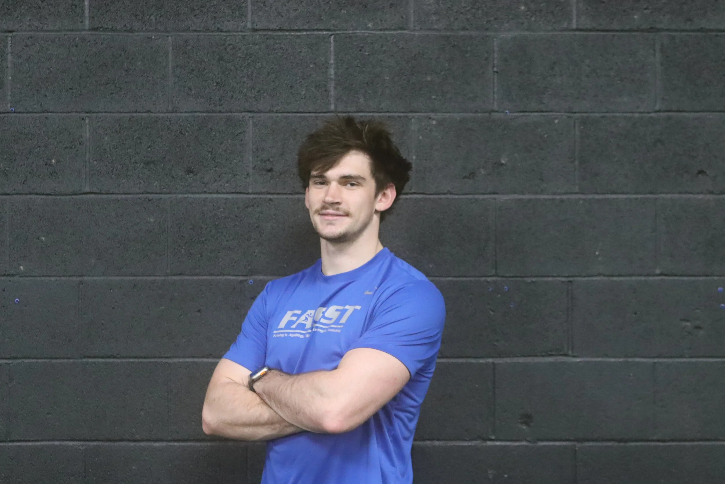 A young man with brown hair and a slight beard, standing with arms crossed, wearing a blue athletic shirt, in front of a dark gray brick wall.