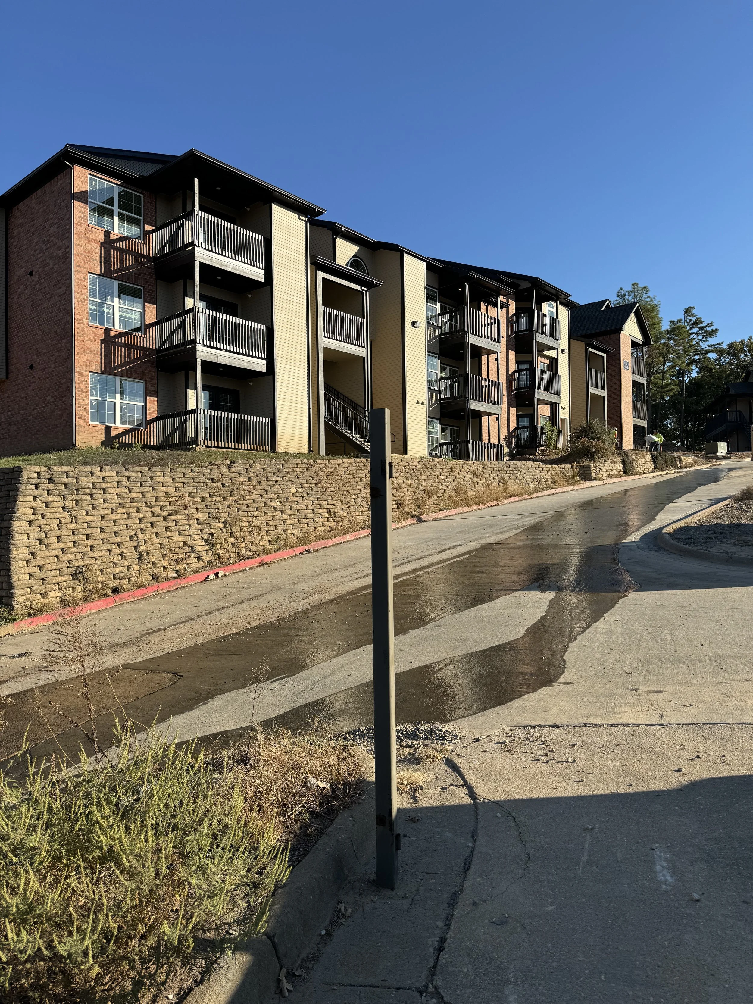 Multi-story residential apartment building with brick and siding exterior, multiple balconies, and an uphill driveway with a curb and a parking barrier in front.