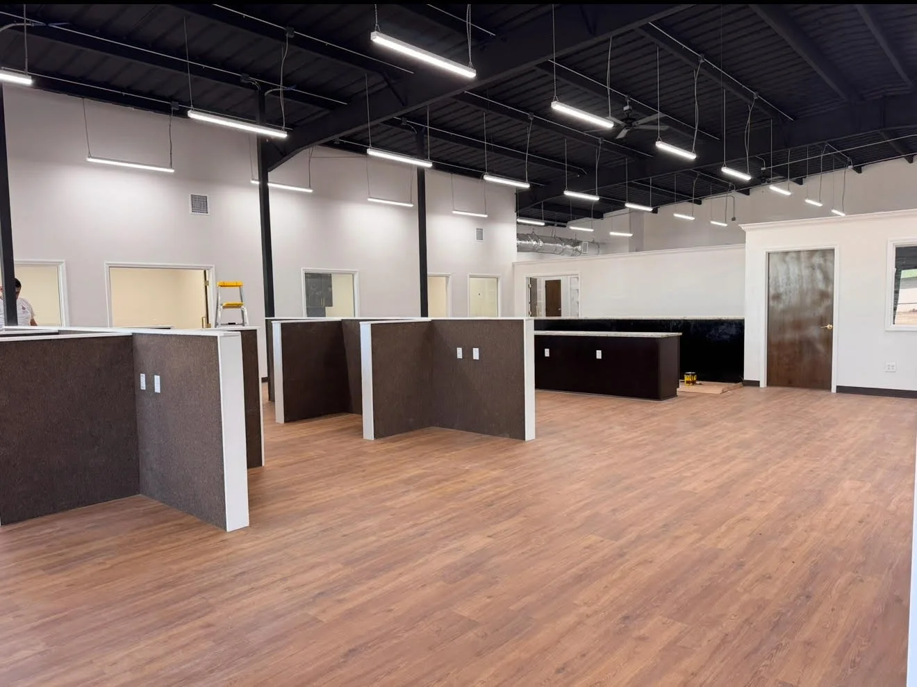 An empty commercial space with dark accent walls, wooden flooring, and multiple workstations separated by brown dividers. Overhead lighting fixtures hang from the ceiling, and several doorways and windows are visible along the walls.
