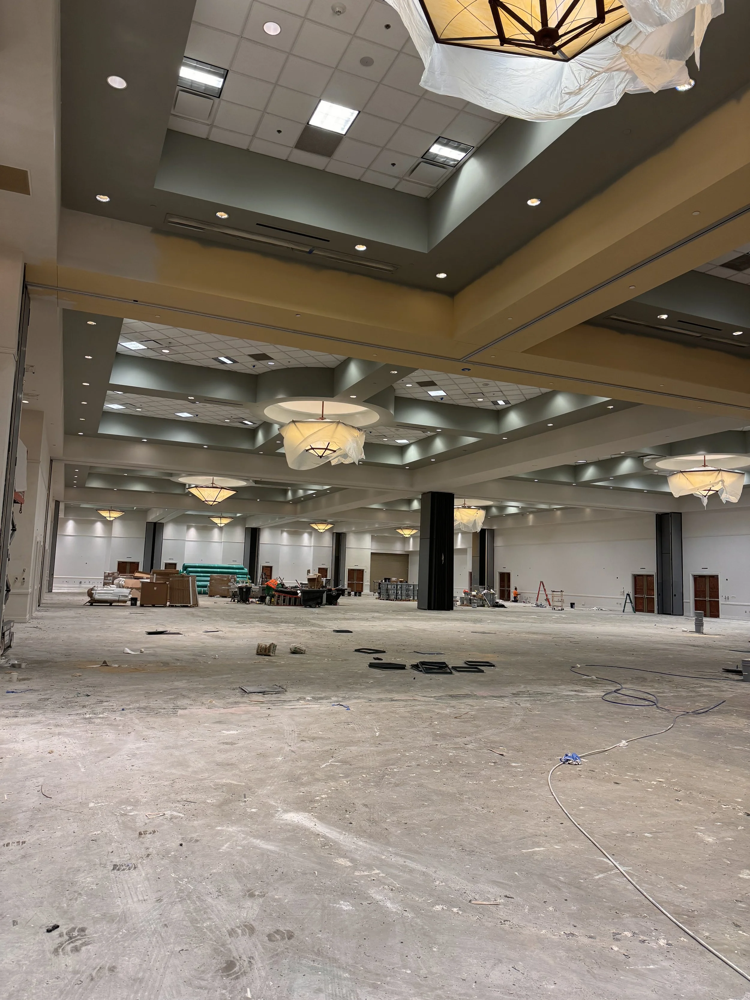 Empty large banquet hall under construction, with suspended light fixtures, construction materials, and tools scattered on the unfinished floor.