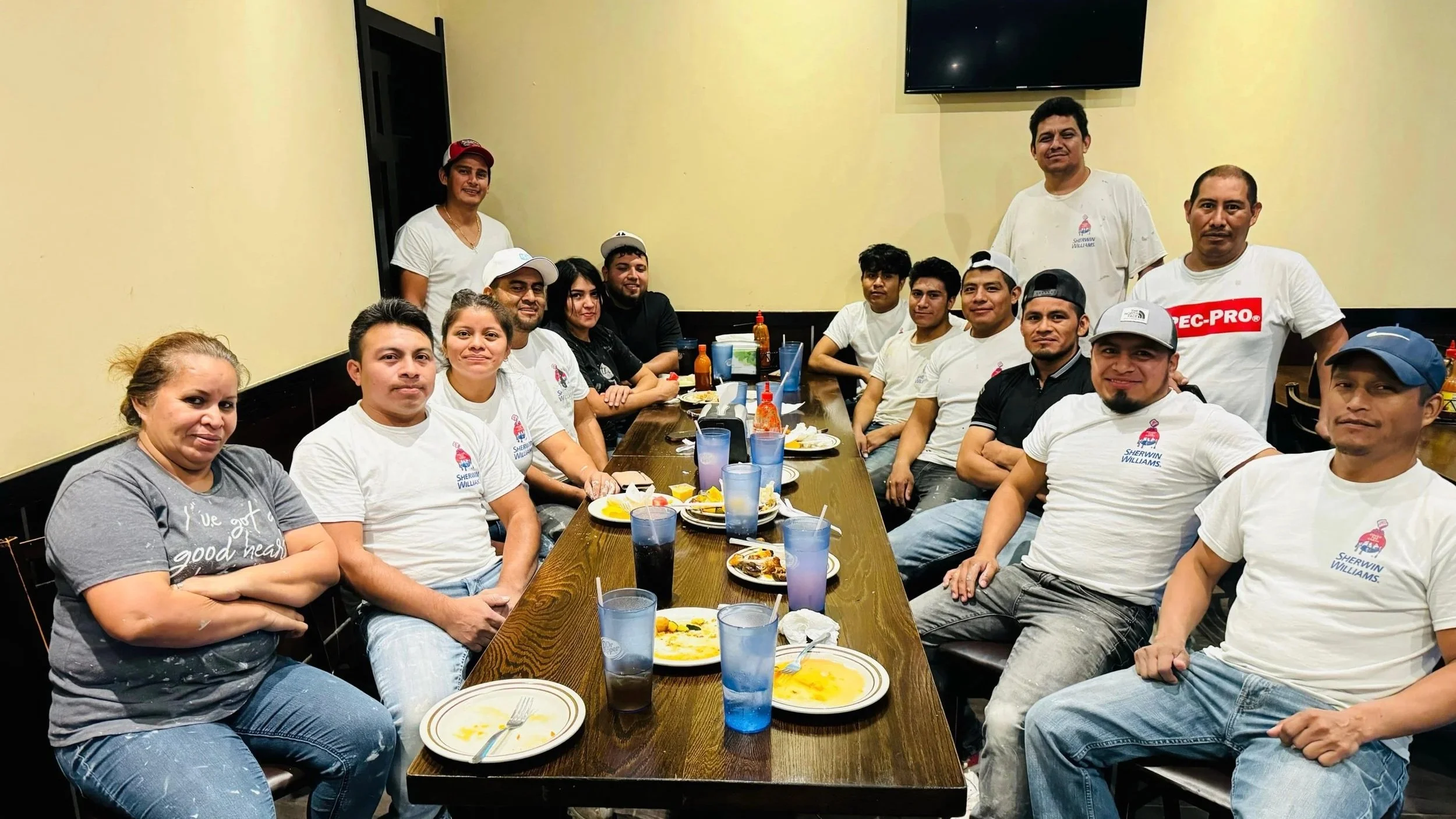 Group of people sitting around a long dining table after a meal, with plates of food and drinks, some people standing and smiling, in a restaurant setting.