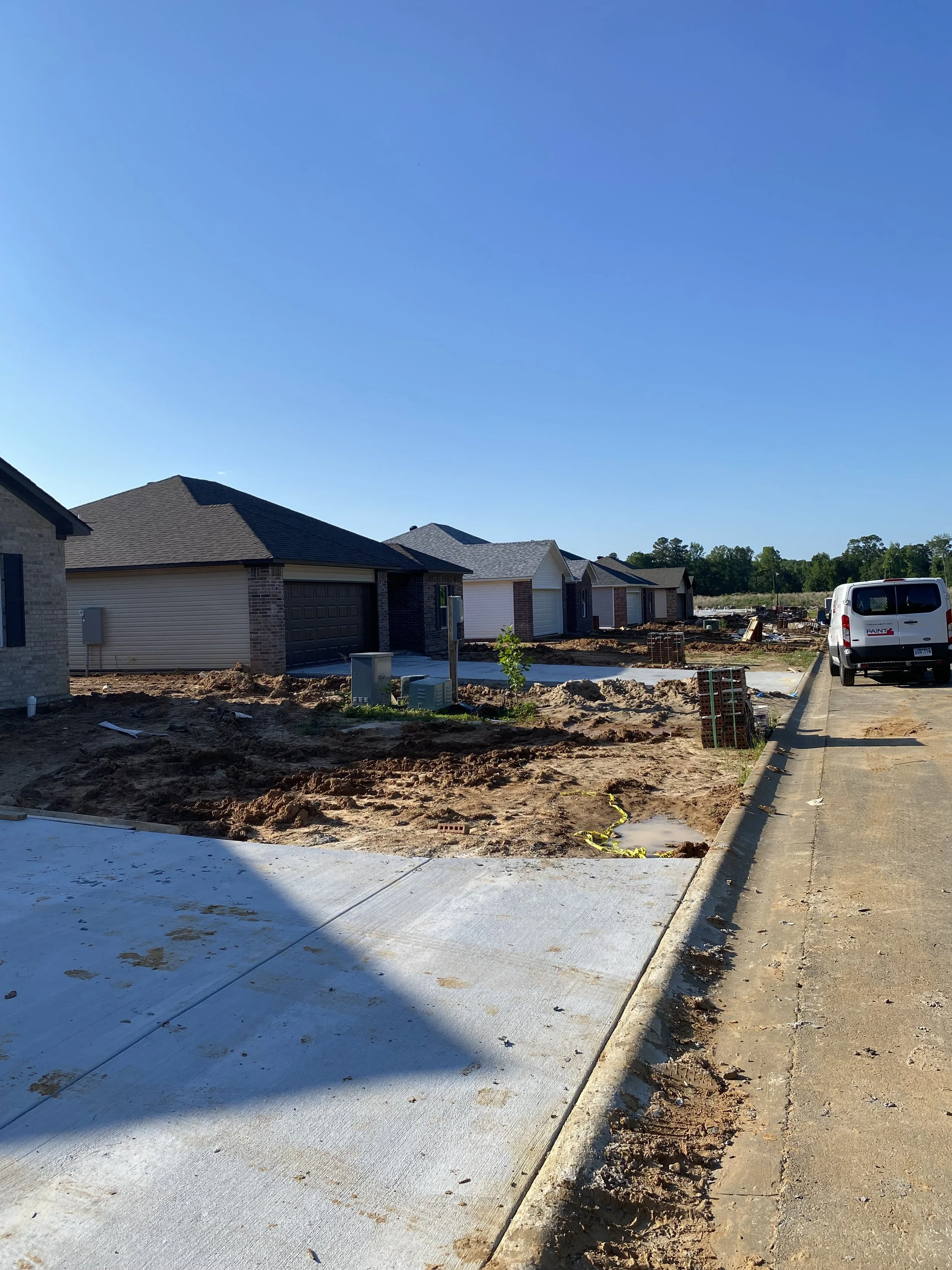 Construction site with newly built houses, a parked white van, a sidewalk, and construction materials under a clear blue sky.