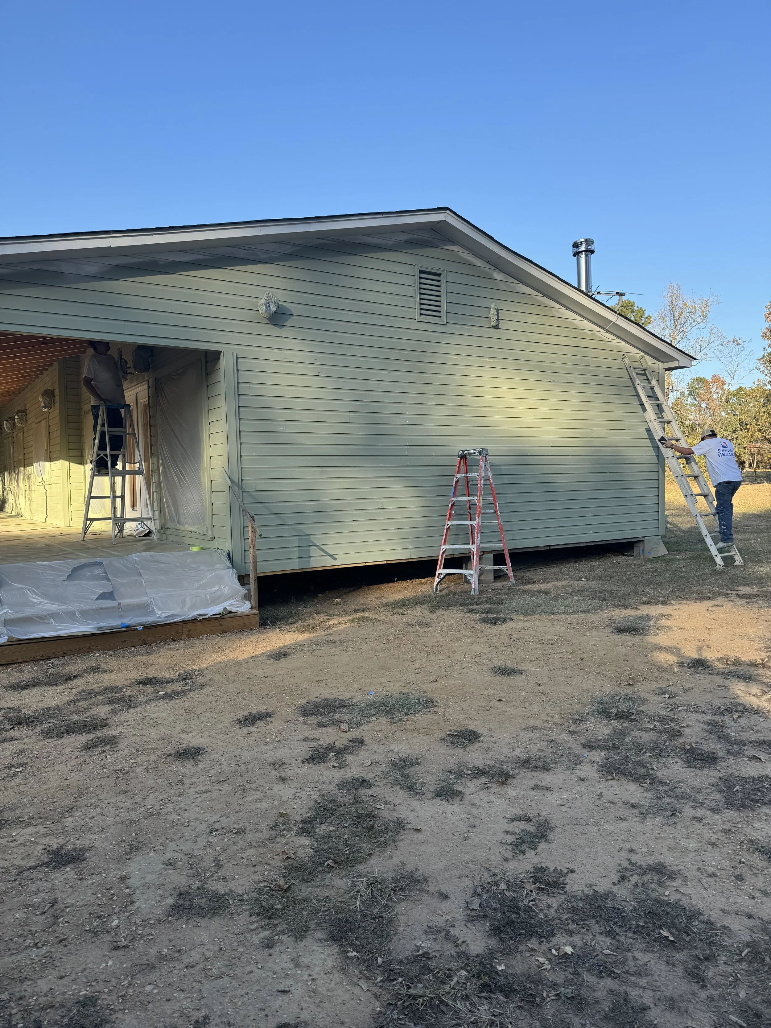 People painting the exterior of a house with siding using ladders and brushes on a sunny day.