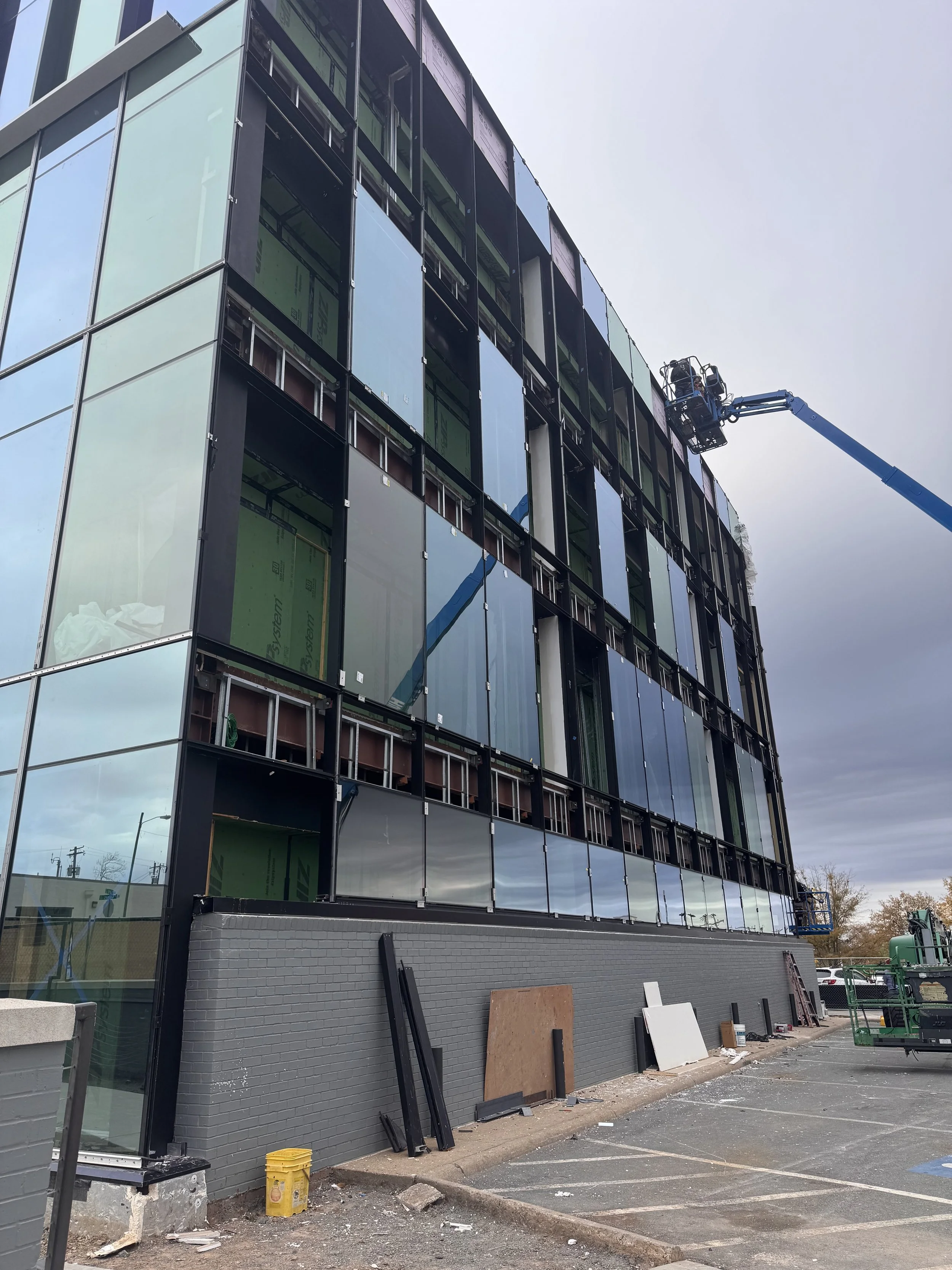 A building under construction with glass panels being installed on the exterior. Two construction lifts are present, one on the right side and one in the upper part of the building. Construction materials and equipment are on the ground nearby.