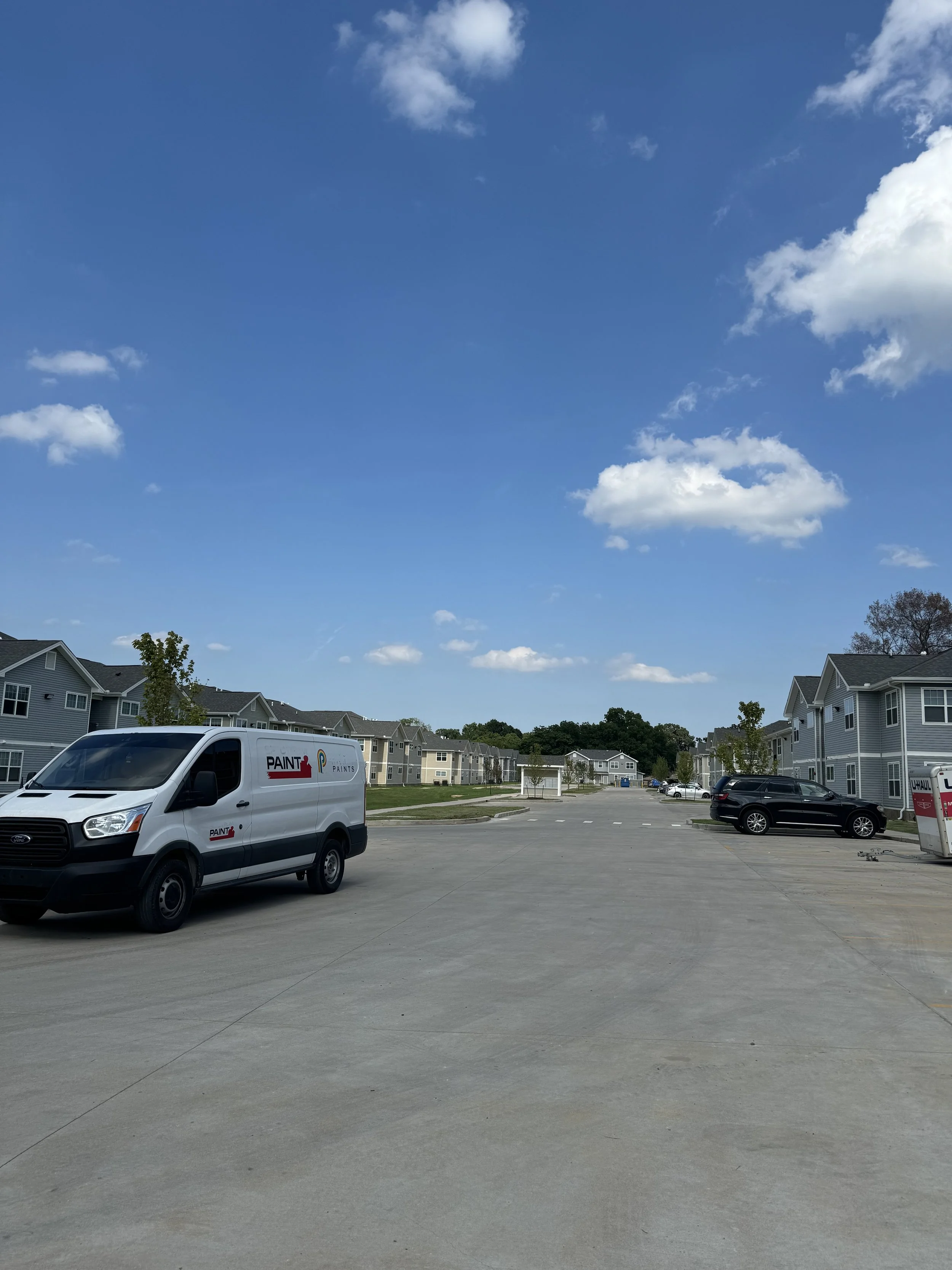 A suburban parking lot with a white van labeled 'Paint' parked on the left, multiple residential houses surrounding the lot, a U-Haul truck on the right, and a bright blue sky with scattered clouds overhead.