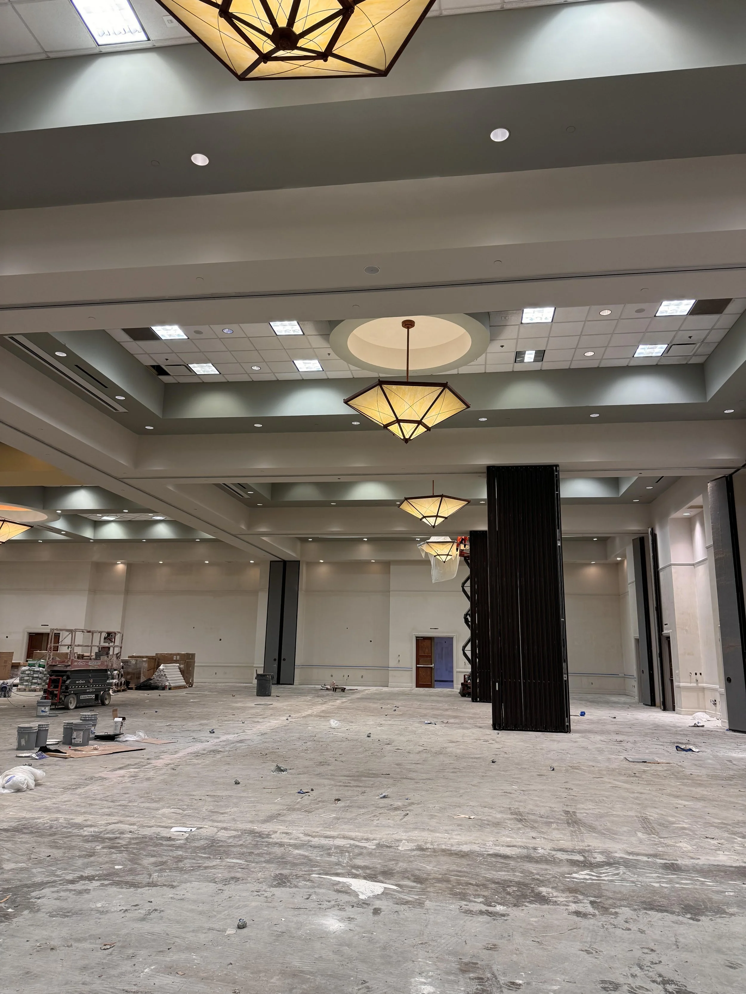 Interior of a large, unfinished event hall or conference room under construction, with high ceilings, modern chandeliers, and construction equipment scattered on the floor.