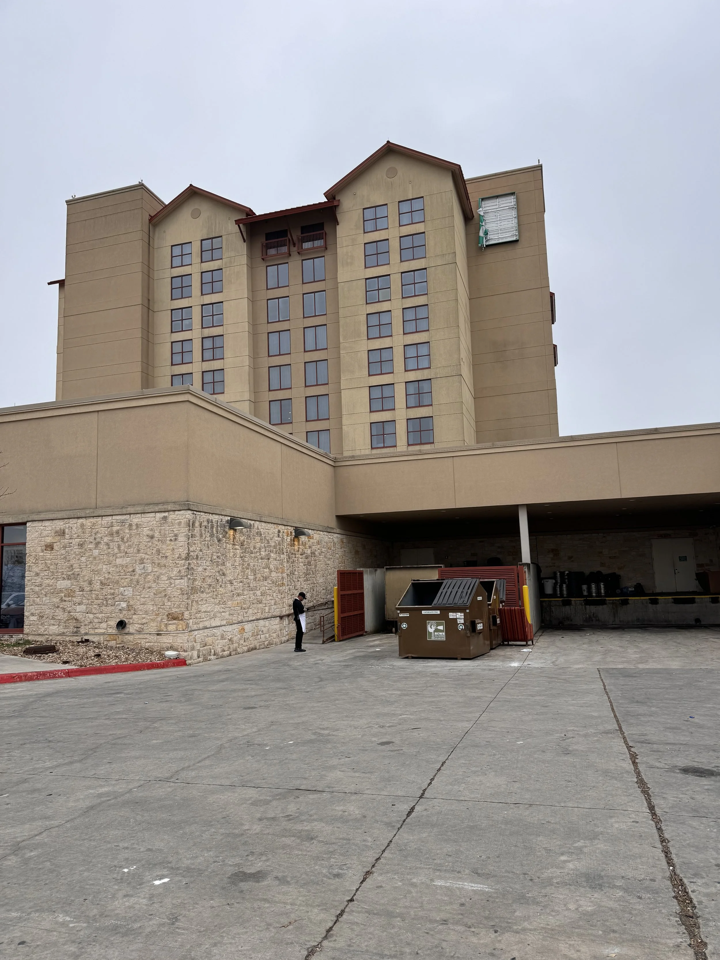 A person standing outside a large beige multi-story building near a dumpster and some recycling bins.