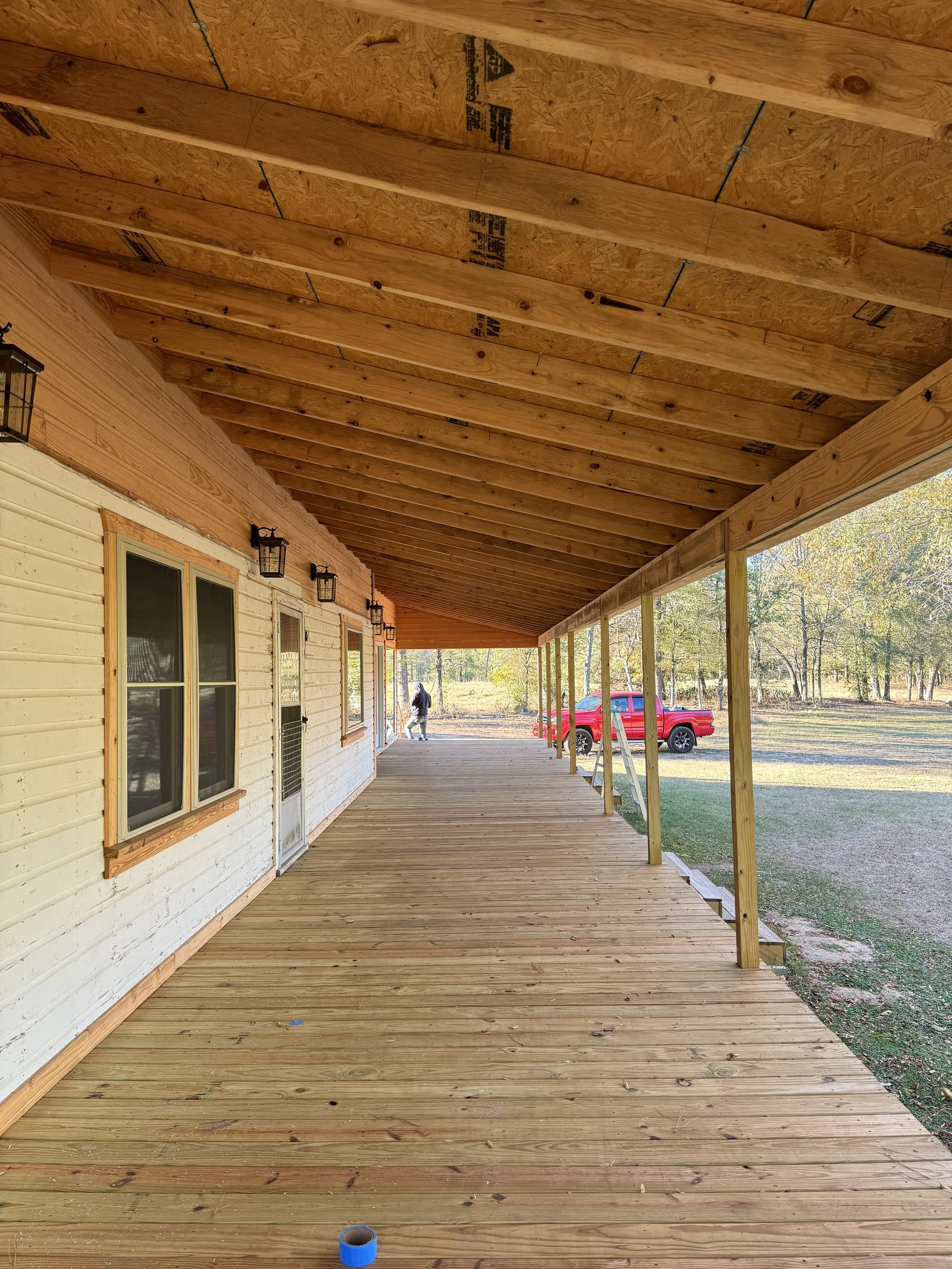 A newly built wooden front porch with a ceiling, railing, and support beams, attached to a house with off-white siding, multiple windows, and outdoor lighting fixtures. A person and a red pickup truck are visible in the background.