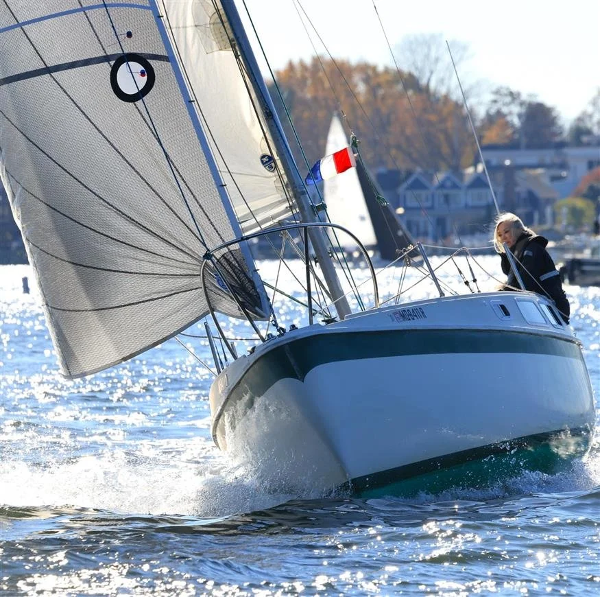 A sailboat on the water with a person on deck, surrounded by houses and trees in the background.