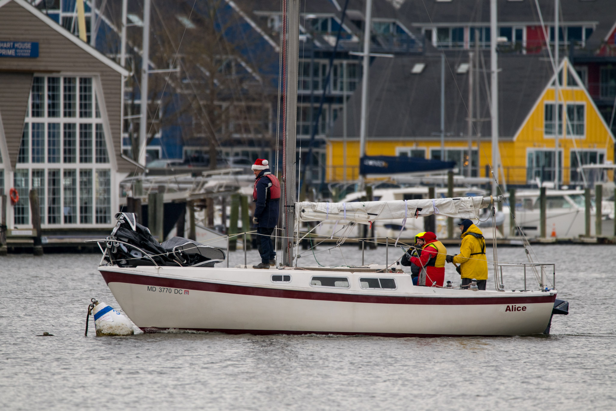 Three people on a sailboat named Alice on a waterfront, with colorful houses and docked boats in the background. One person is standing, and two are seated near the stern, all dressed in jackets and hats.