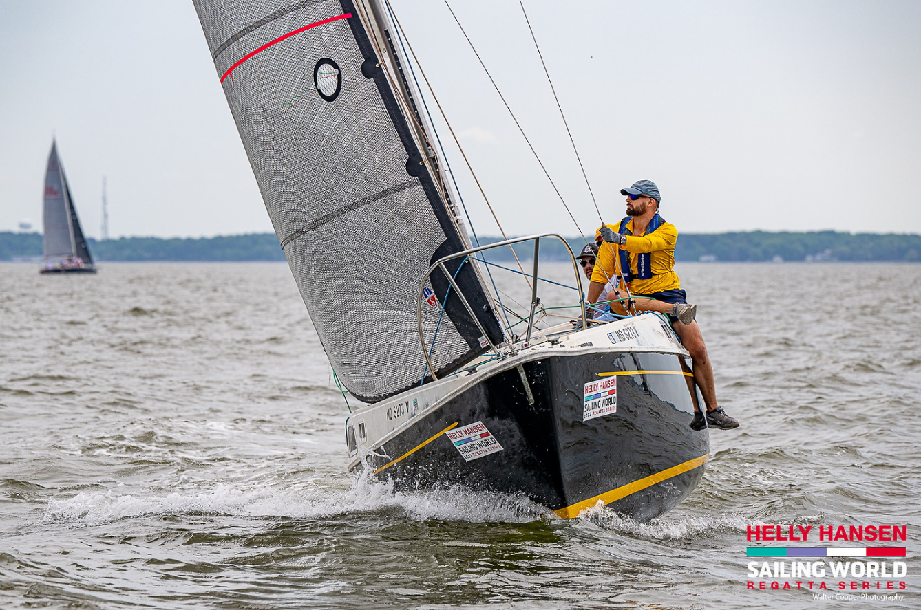 Two sailors on a sailboat leaning out over the edge in a regatta, with one man in a yellow jacket and blue cap steering, on a choppy body of water with other sailboats in the background.