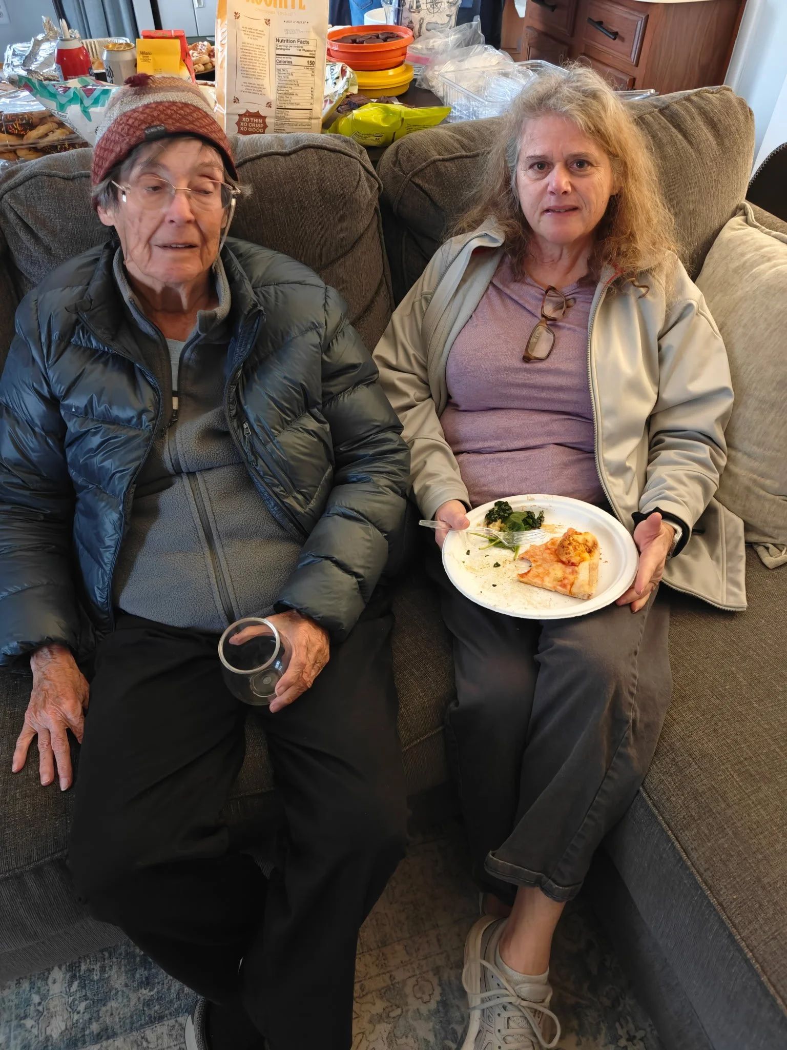 Two women sitting on a sofa in a living room, one holding a plate with partially eaten food and the other with a glass in hand, in front of a cluttered table with snacks and food packages.