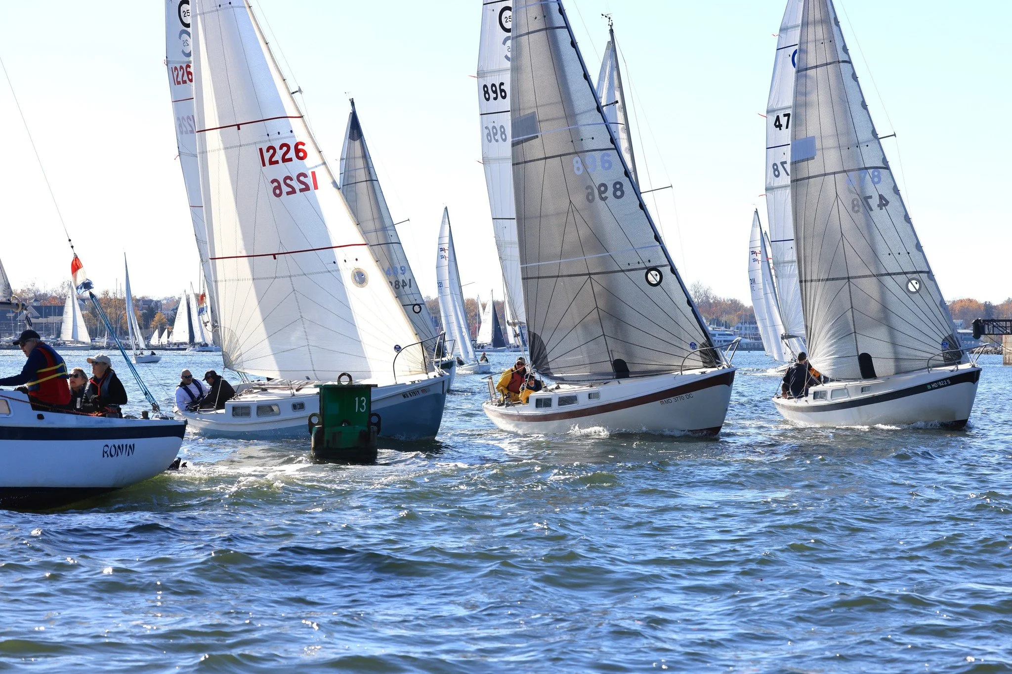 Sailing race with multiple sailboats on the water during daytime, with crew members on deck and sails catching the wind.