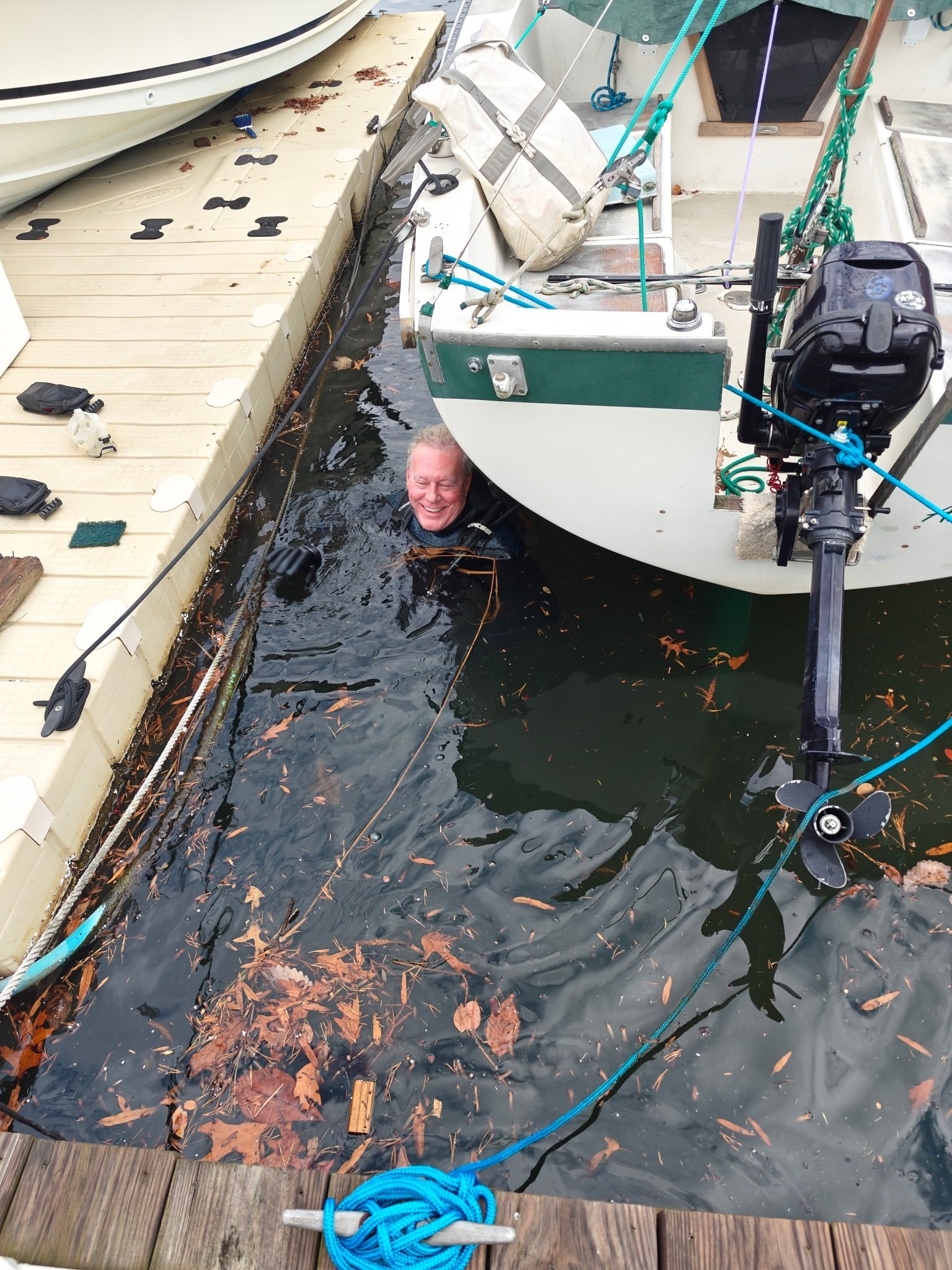 A man is smiling and floating in the water next to a docked boat, holding onto the dock, with fallen leaves floating on the water's surface.