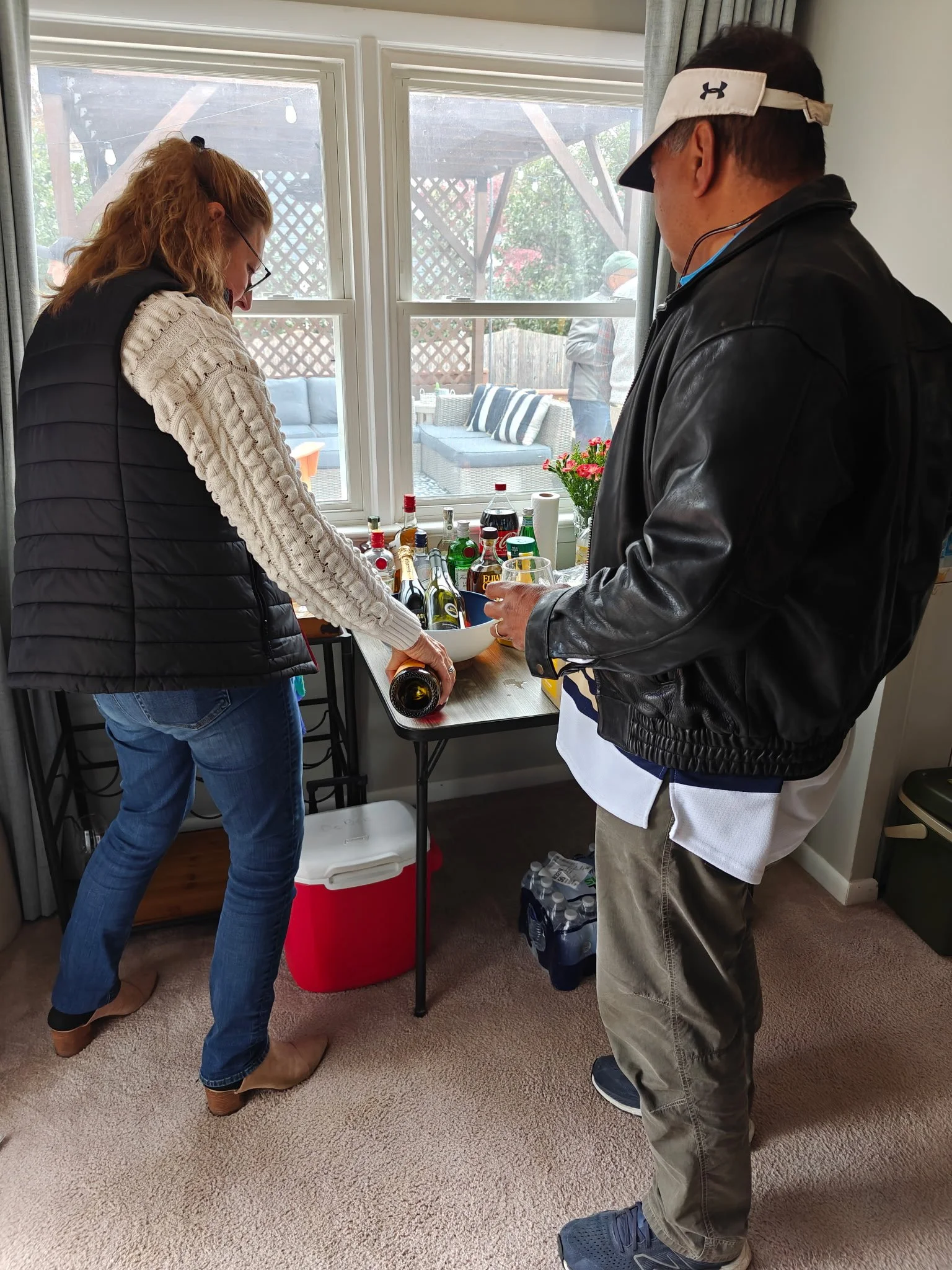 Two people standing near a window, preparing drinks with bottles and mixers on a table, with an outdoor patio visible through the window.
