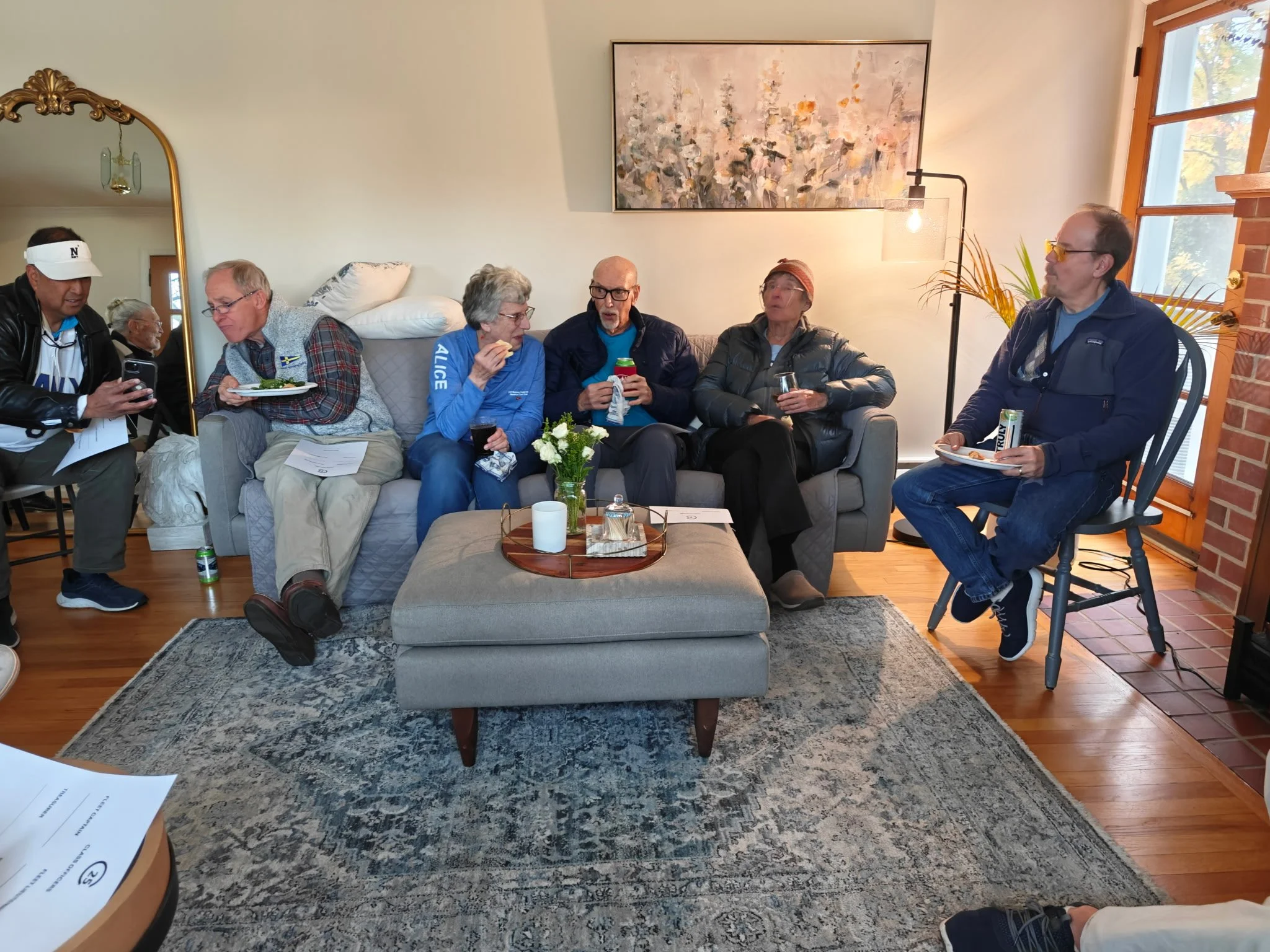 Six people sitting and standing in a living room, some holding food and drinks, engaged in conversation. The room features a gray sofa, wooden flooring, a round coffee table with a flower arrangement, large window with plants, and a modern painting o