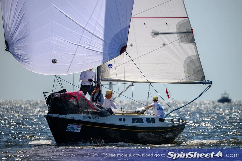 People sailing on a small black and white sailboat with large white sails on a sunny day with calm ocean waters; a large cruise ship is visible in the distance.