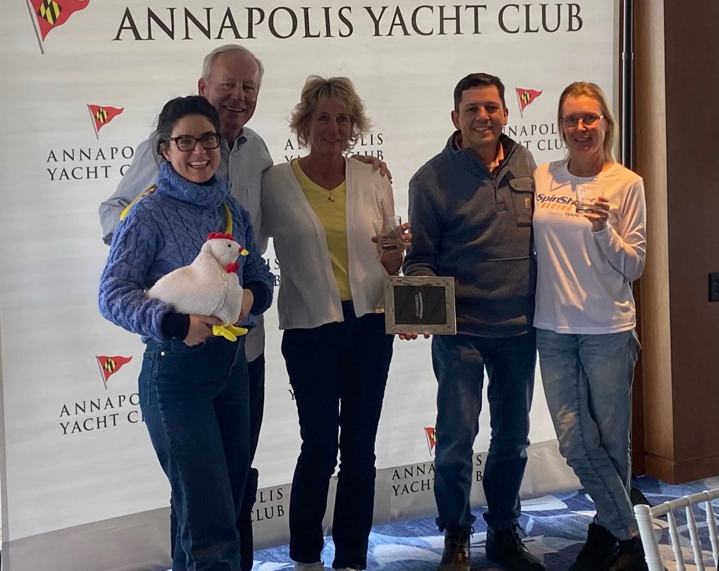 Group of five people standing in front of a backdrop reading 'Annapolis Yacht Club,' with one person holding a plush chicken toy and another holding a glass, celebrating or recognizing an achievement.